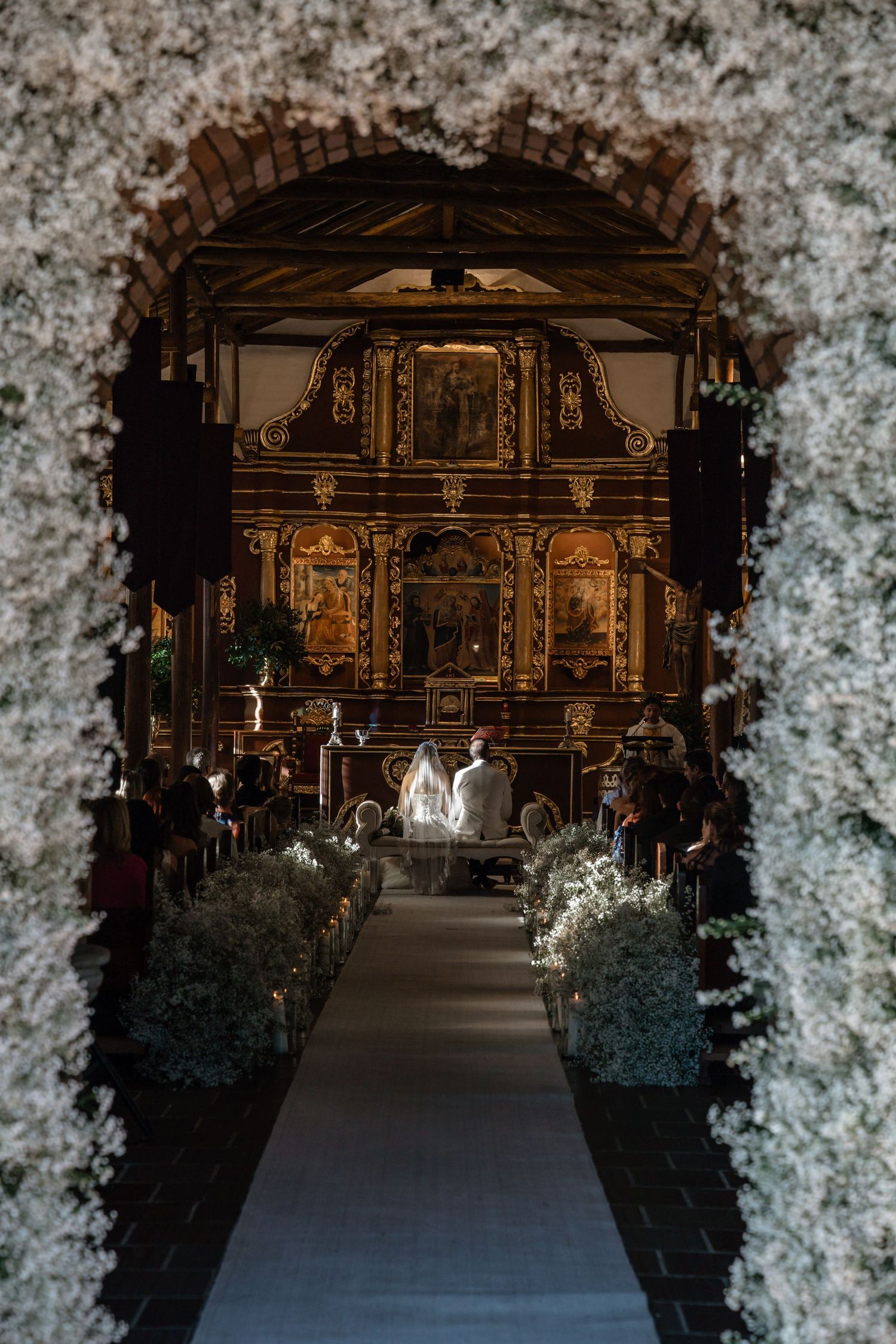 Wedding ceremony viewed through a floral arch in an ornate historic church with golden altar.