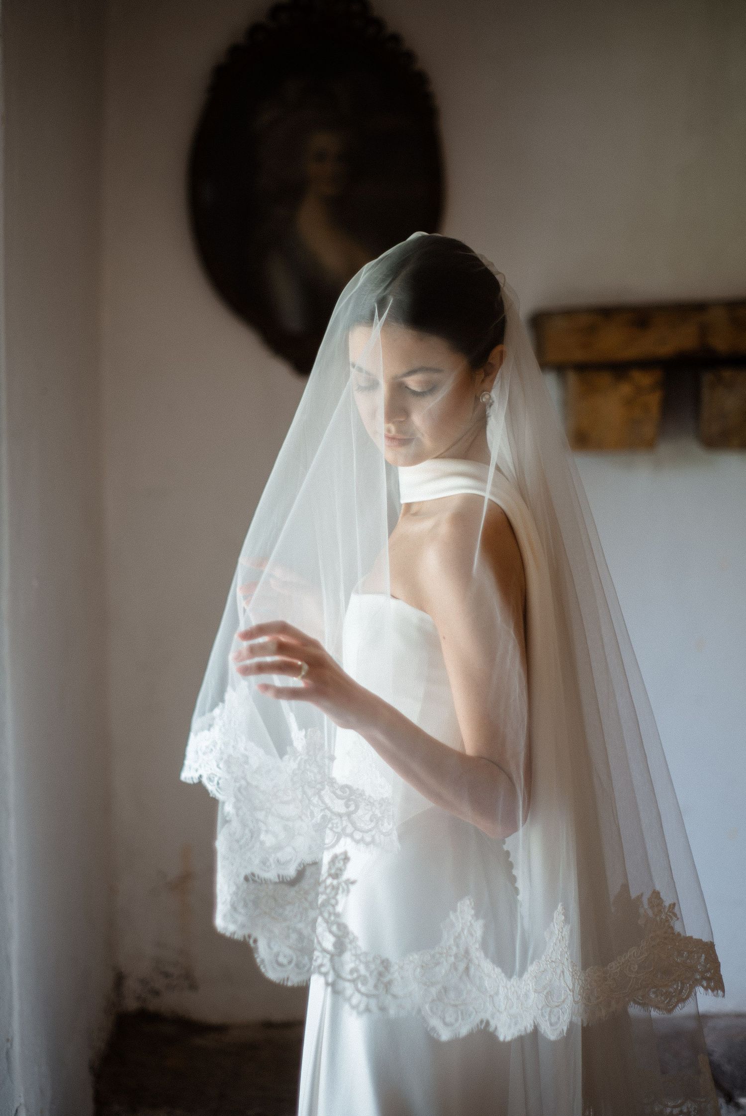 Bride in a strapless white gown wearing a lace-trimmed cathedral veil, gazing down in a vintage interior.