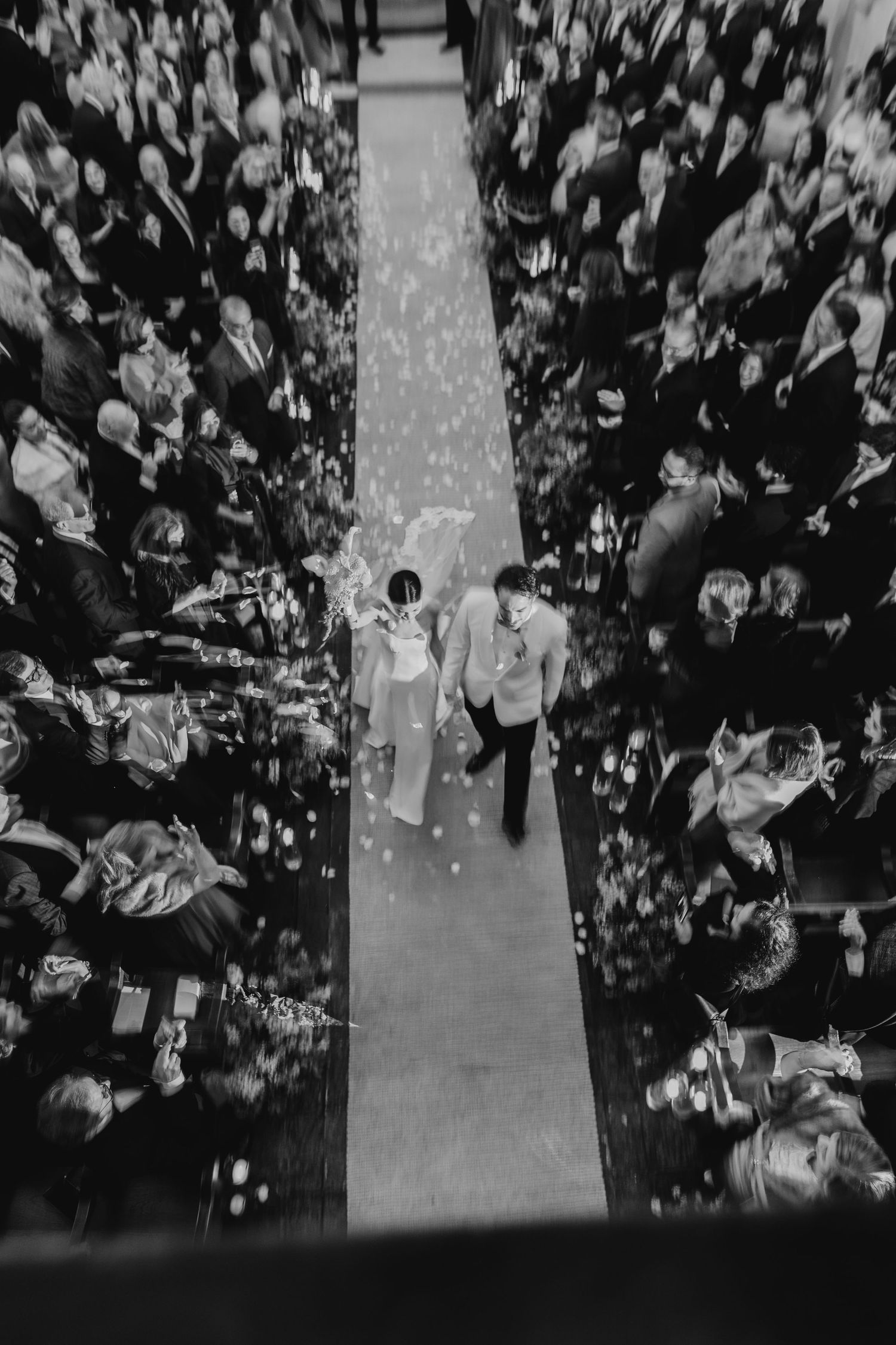 Aerial black and white photo of bride and groom walking down aisle as guests toss confetti.