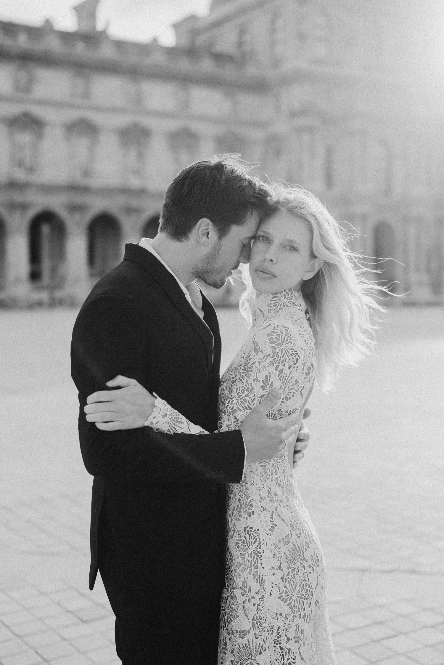 Elegant couple embracing in black and white photo at a grand Parisian courtyard.
