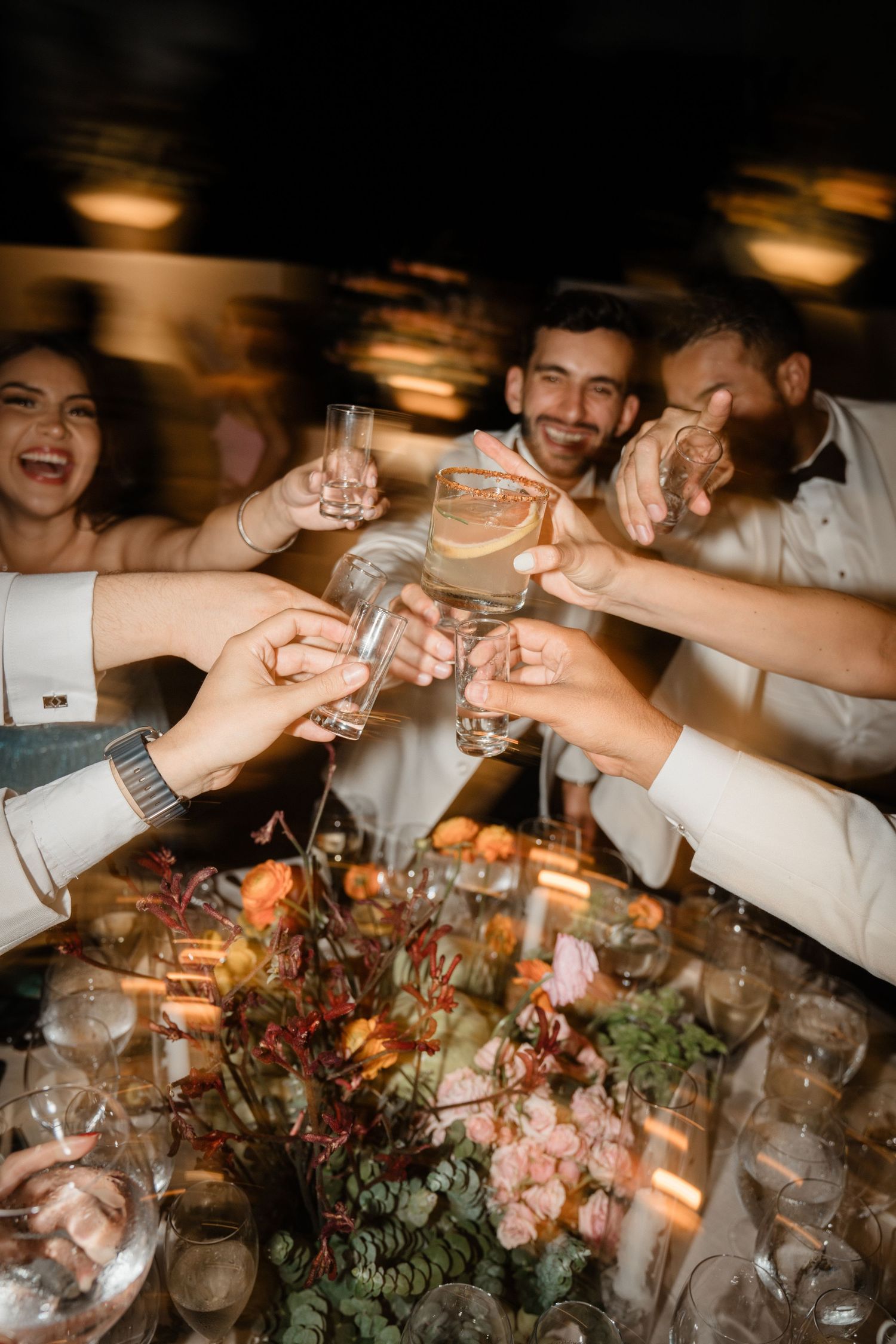 Wedding guests clinking shot glasses in a joyful toast over a floral centerpiece at a reception dinner.