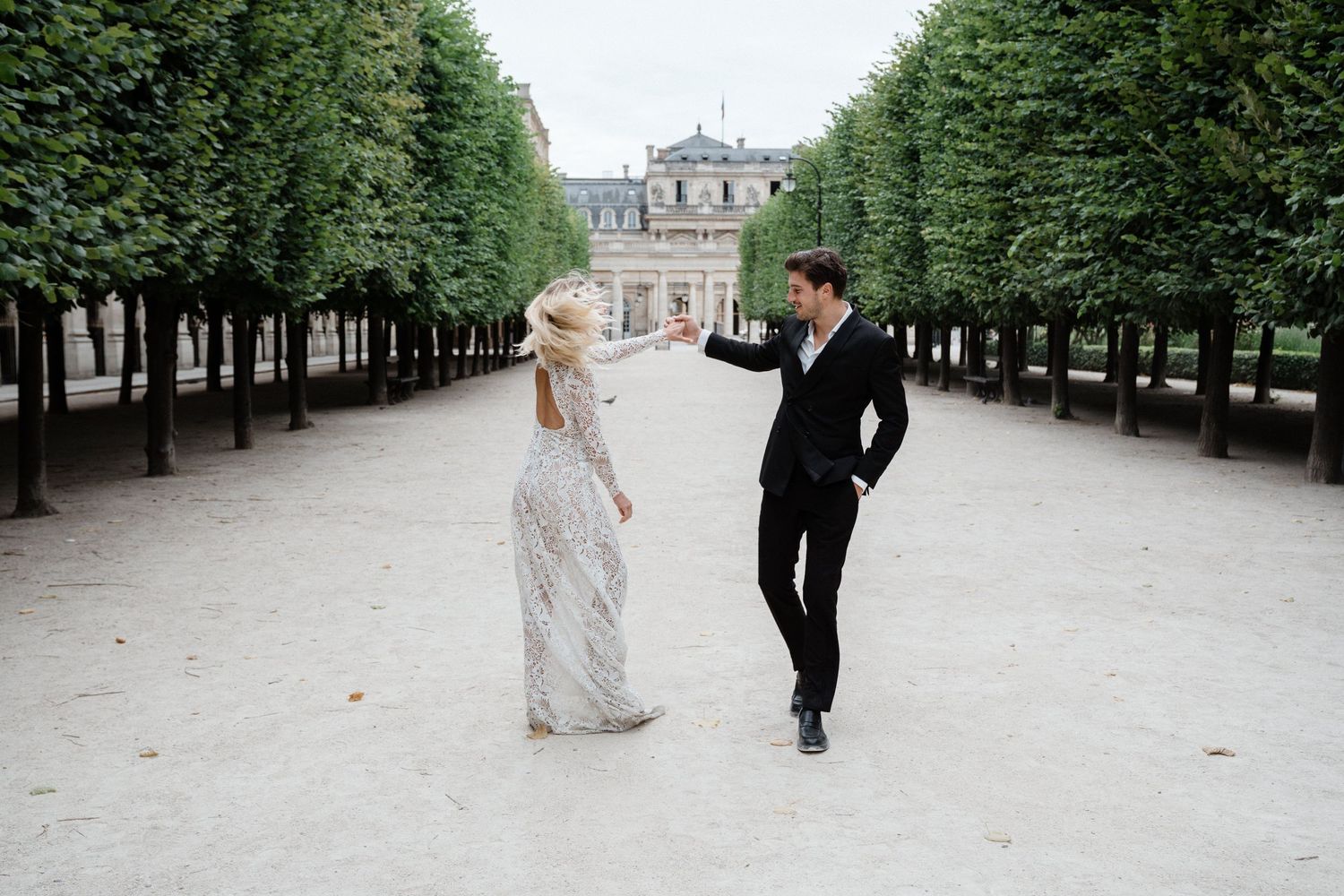 Couple dancing in a Parisian garden alley with trimmed trees and a classic building backdrop.