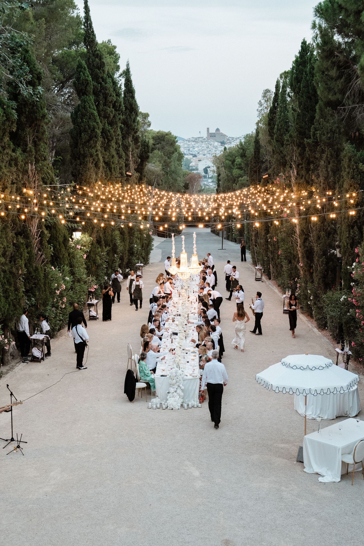 Aerial view of an outdoor wedding reception with string lights lining a long dining table among tall trees.