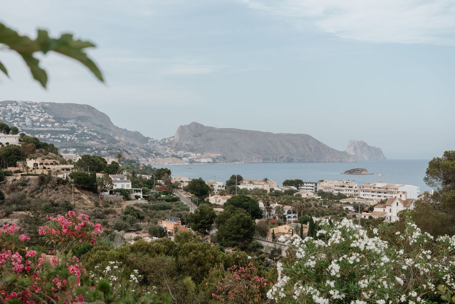 Scenic coastal view with lush flowering gardens, hillside villas, and a calm blue Mediterranean bay.