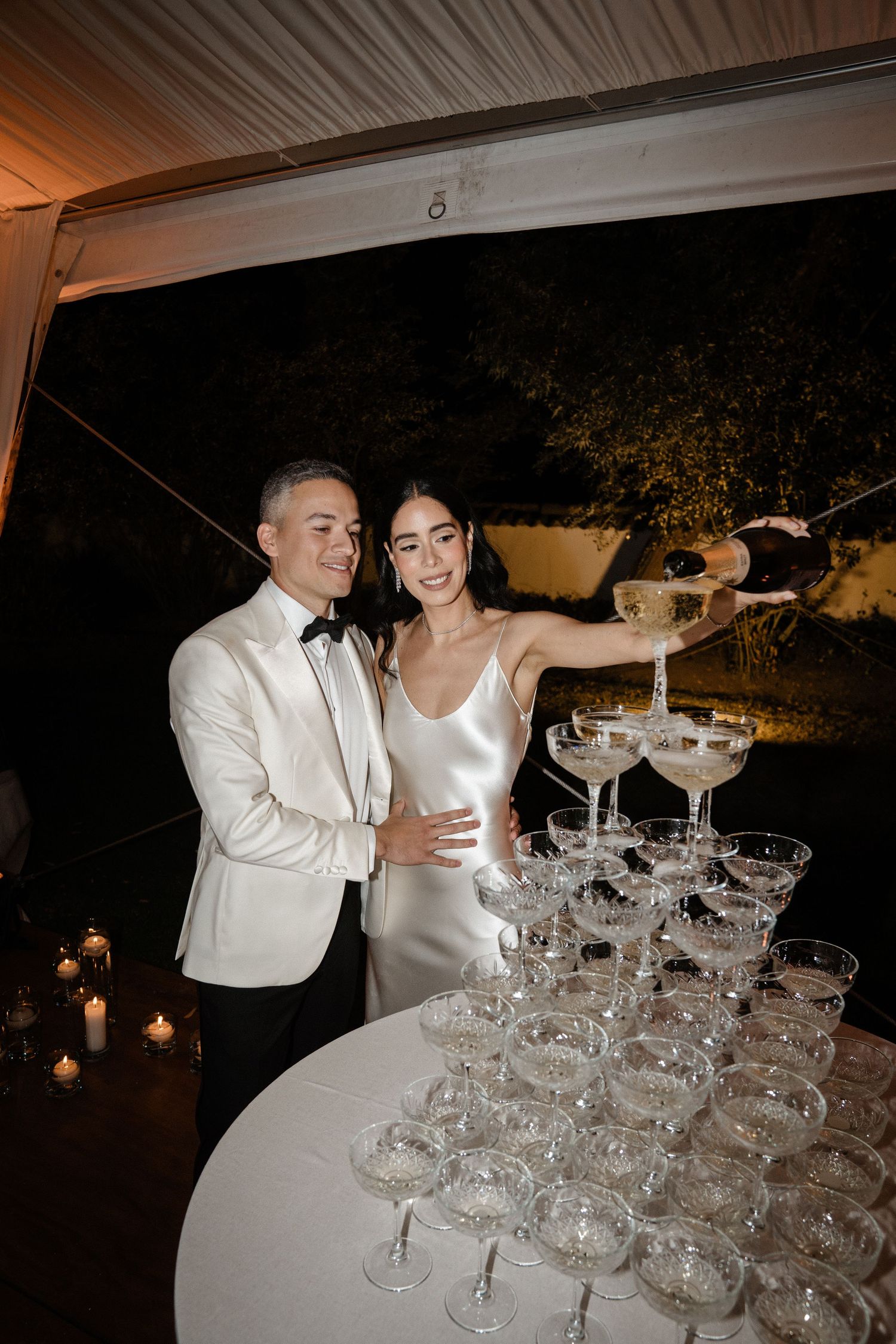 Bride and groom pouring champagne tower at wedding reception at night, wearing white gown and tuxedo.
