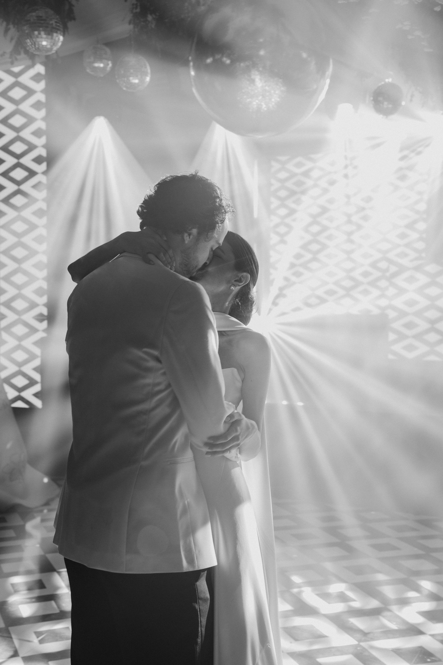 Couple sharing a tender first dance embrace under glowing light rays in a black and white wedding portrait.