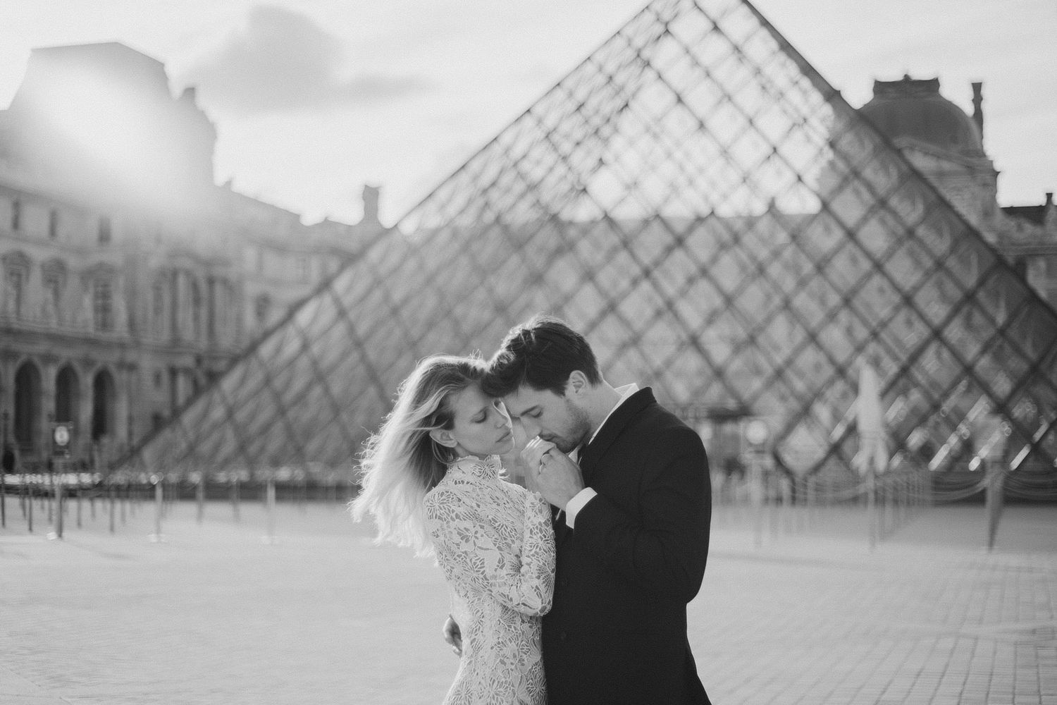 Black and white photo of a couple embracing before the Louvre pyramid in Paris.