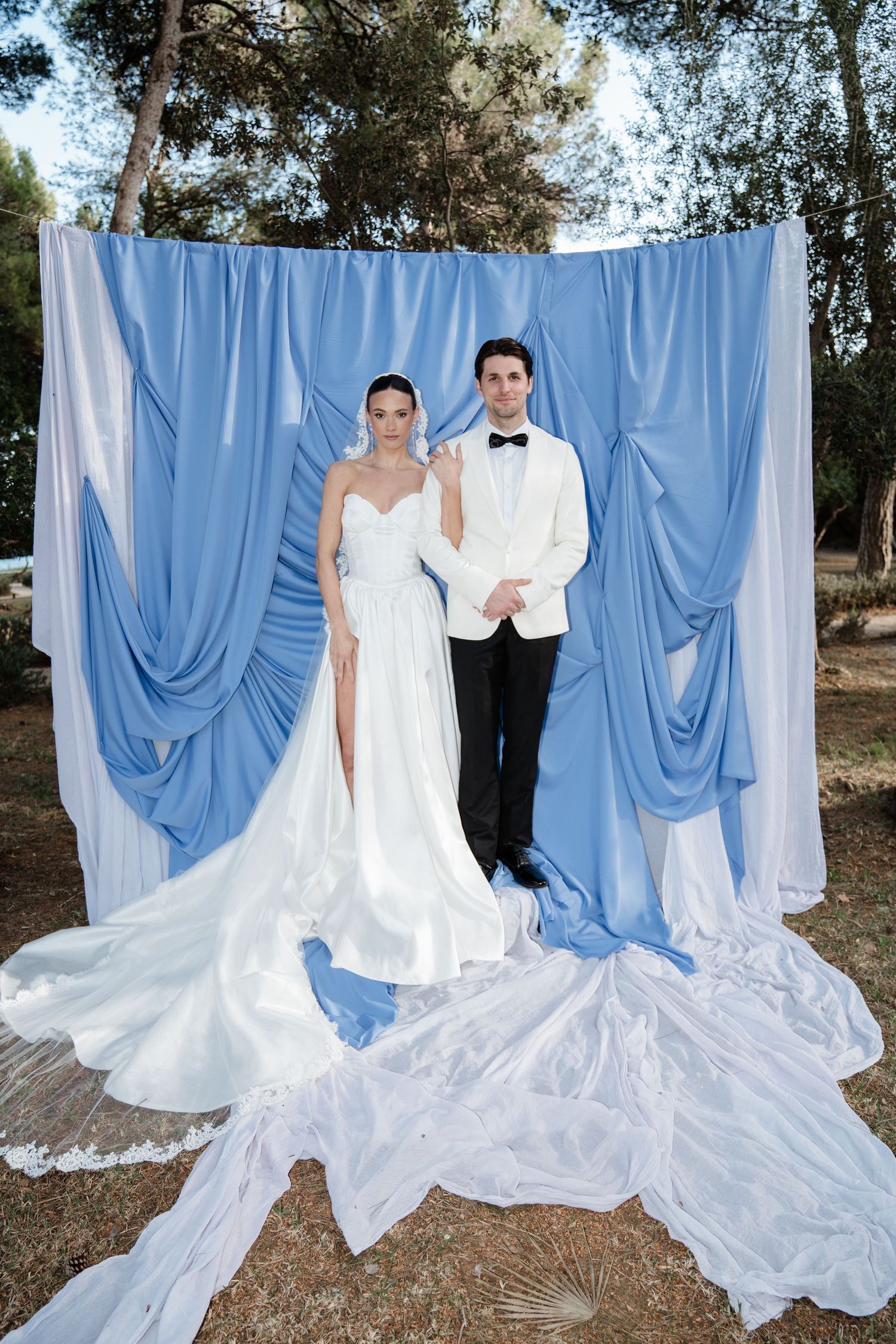 Bride and groom pose before a dramatic blue and white draped fabric backdrop in an outdoor forest setting.