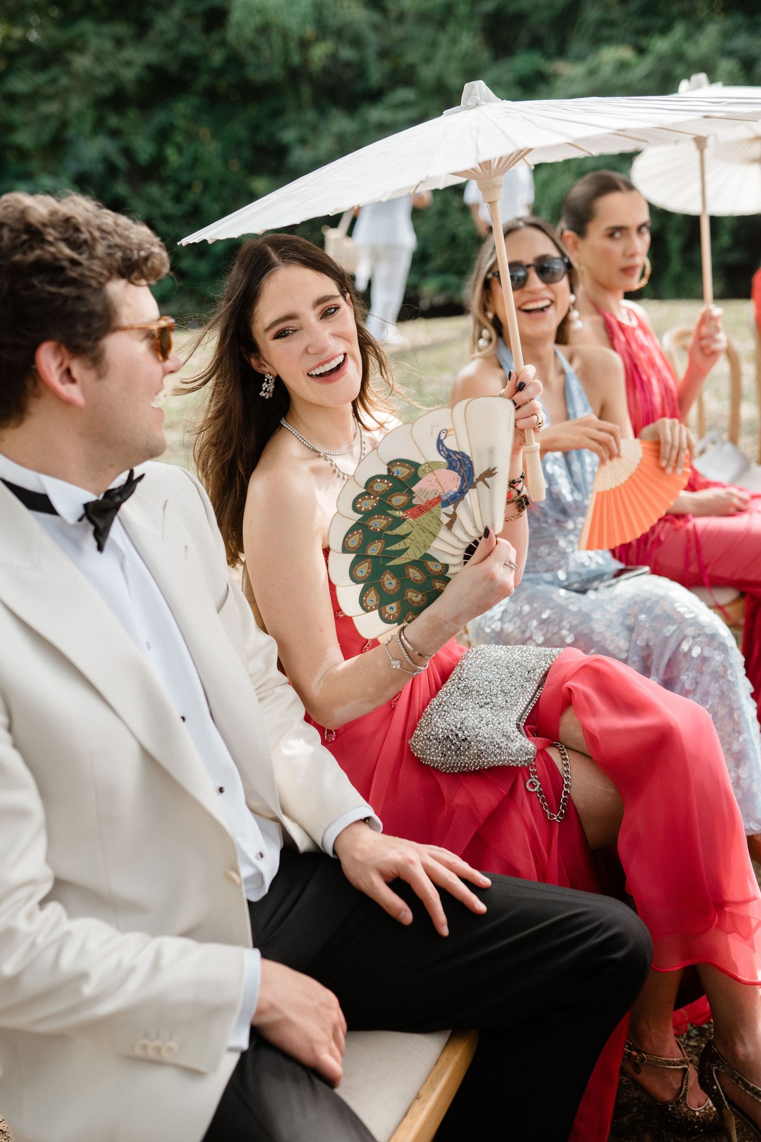Stylish wedding guests laughing with parasols and colorful fans at outdoor summer ceremony.