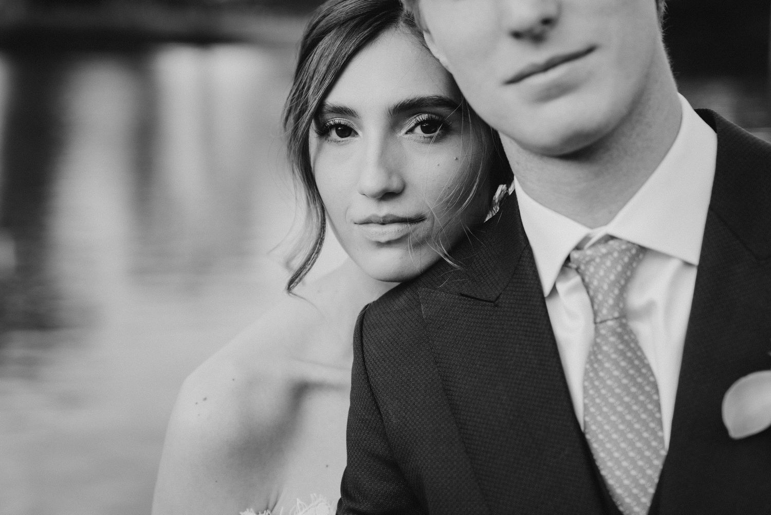Black and white portrait of a bride and groom posing closely together near a reflective waterfront.