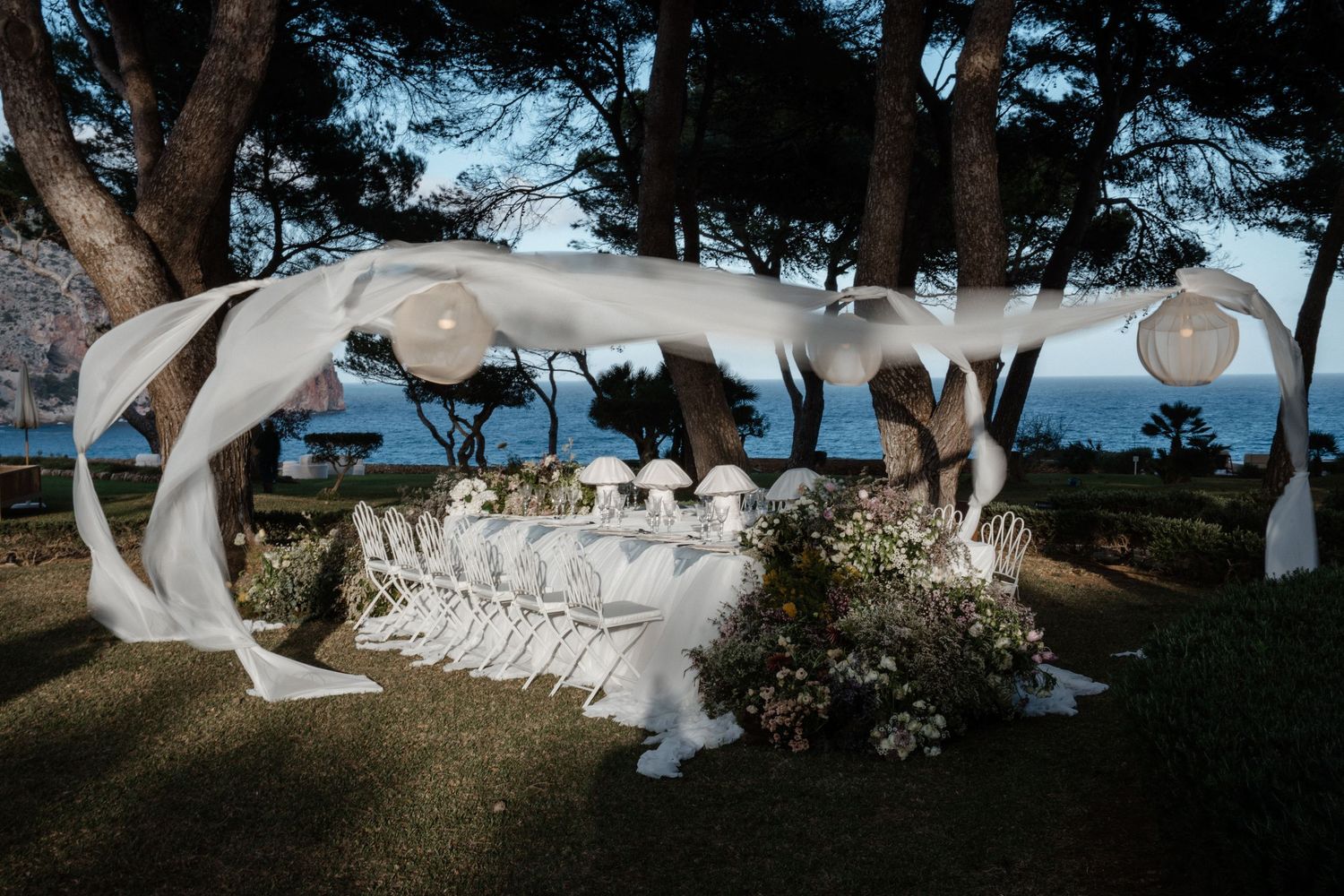 Romantic outdoor evening wedding reception table draped in white fabric beneath pine trees by the sea.