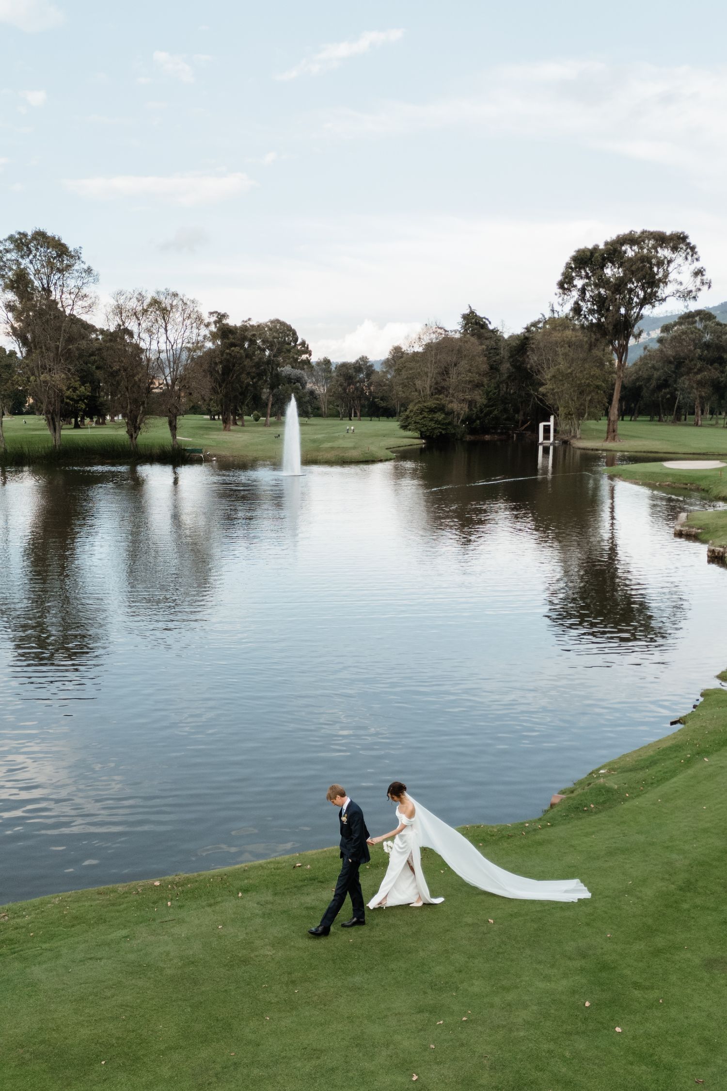 Bride and groom walk hand in hand along a lush green golf course beside a serene pond.