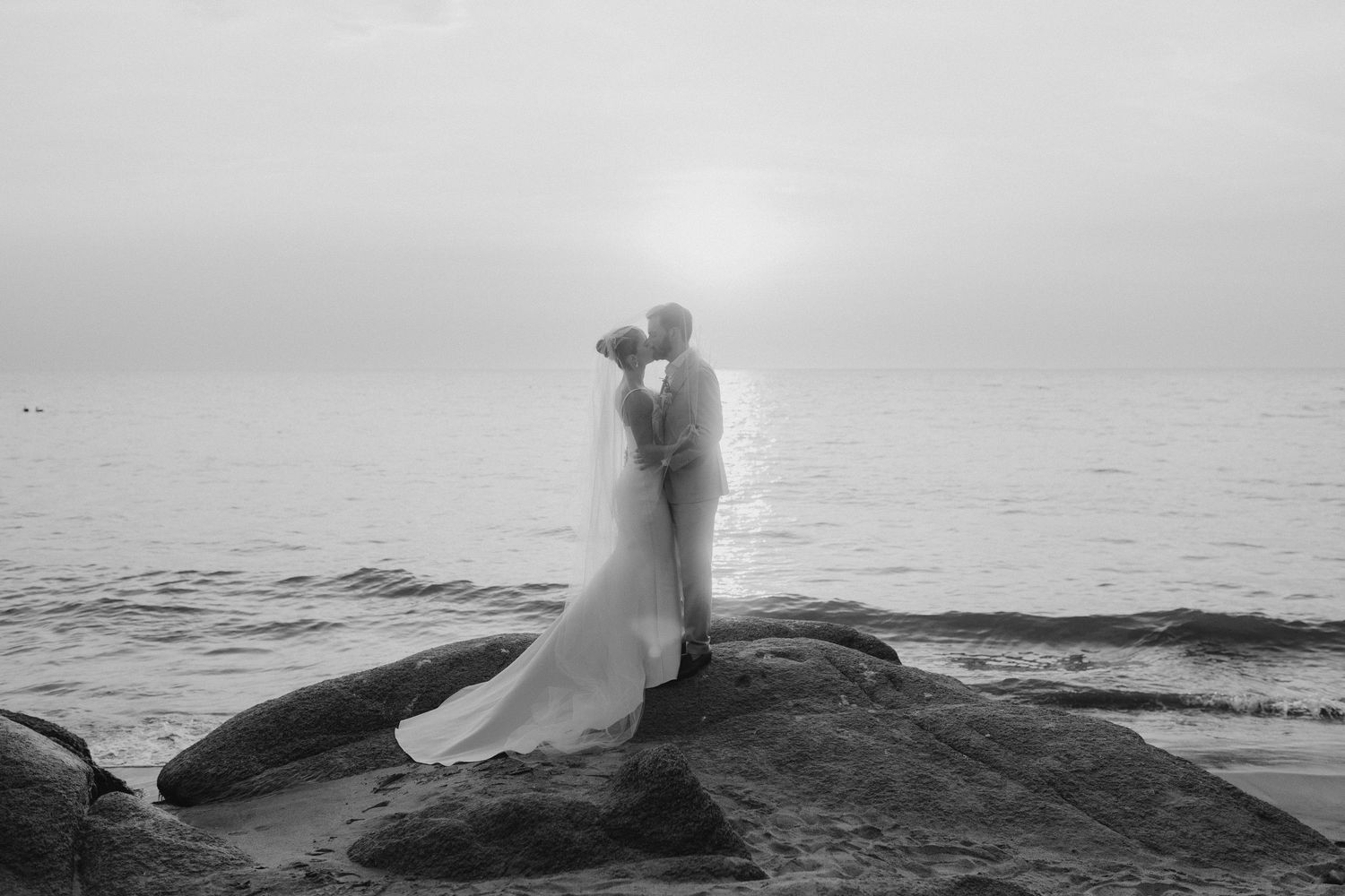 Black and white photo of a bride and groom kissing on rocky shoreline at sunset during their beach wedding.