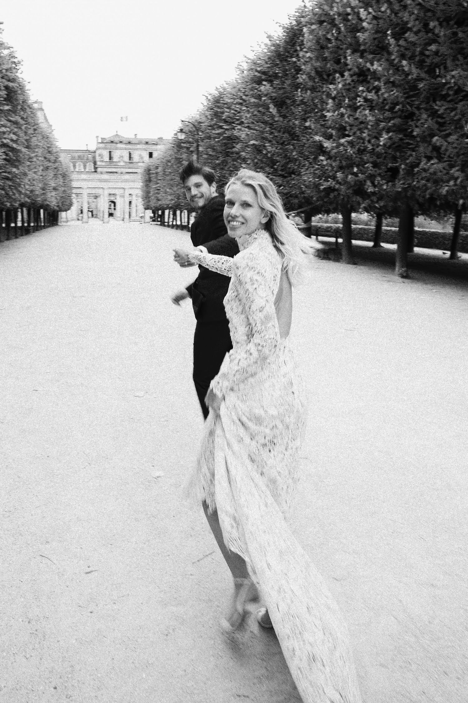 Black and white photo of a couple in formal wear walking through a tree-lined garden path, woman in lace gown glancing back.