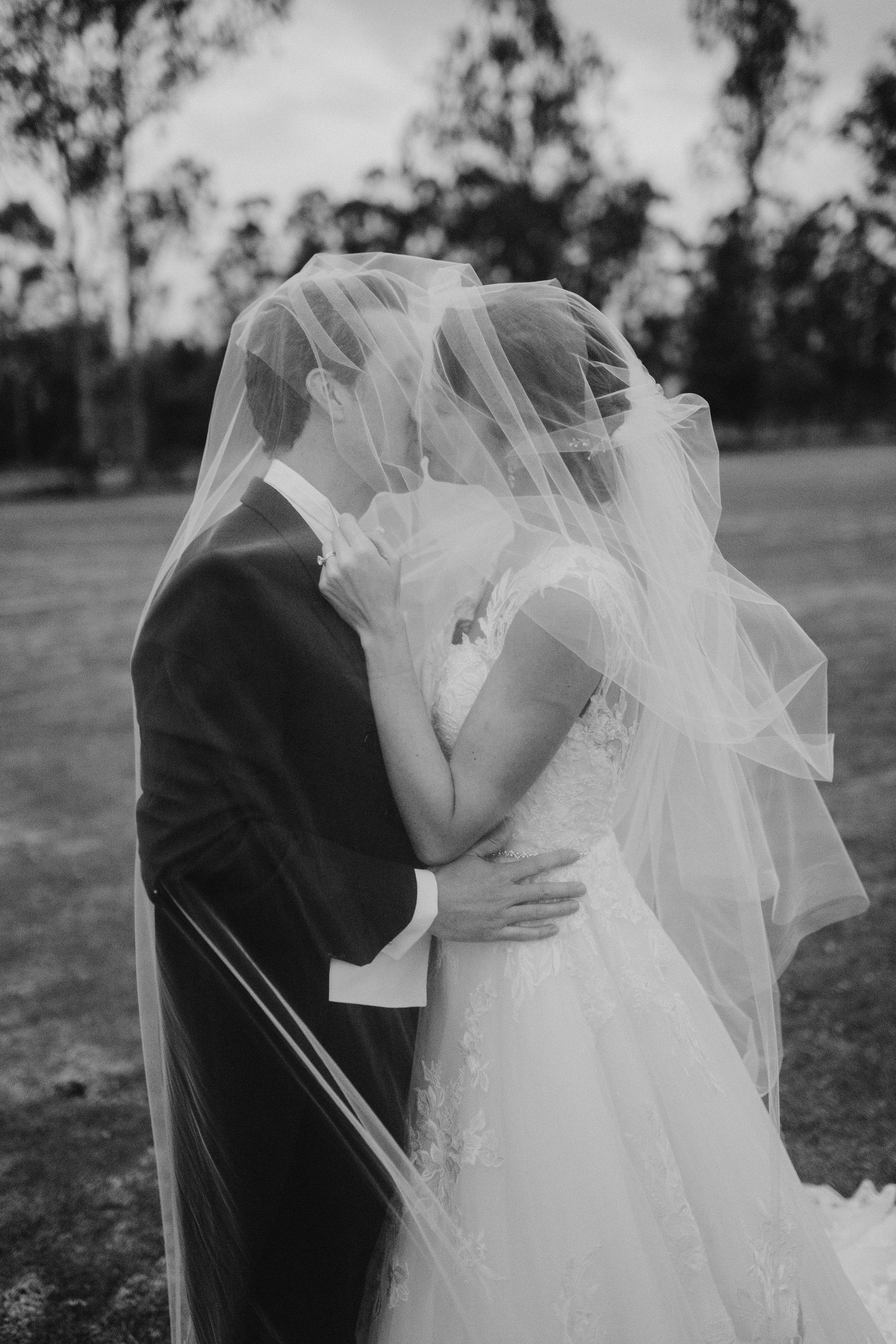 Bride and groom share a kiss hidden beneath a flowing veil in black and white.