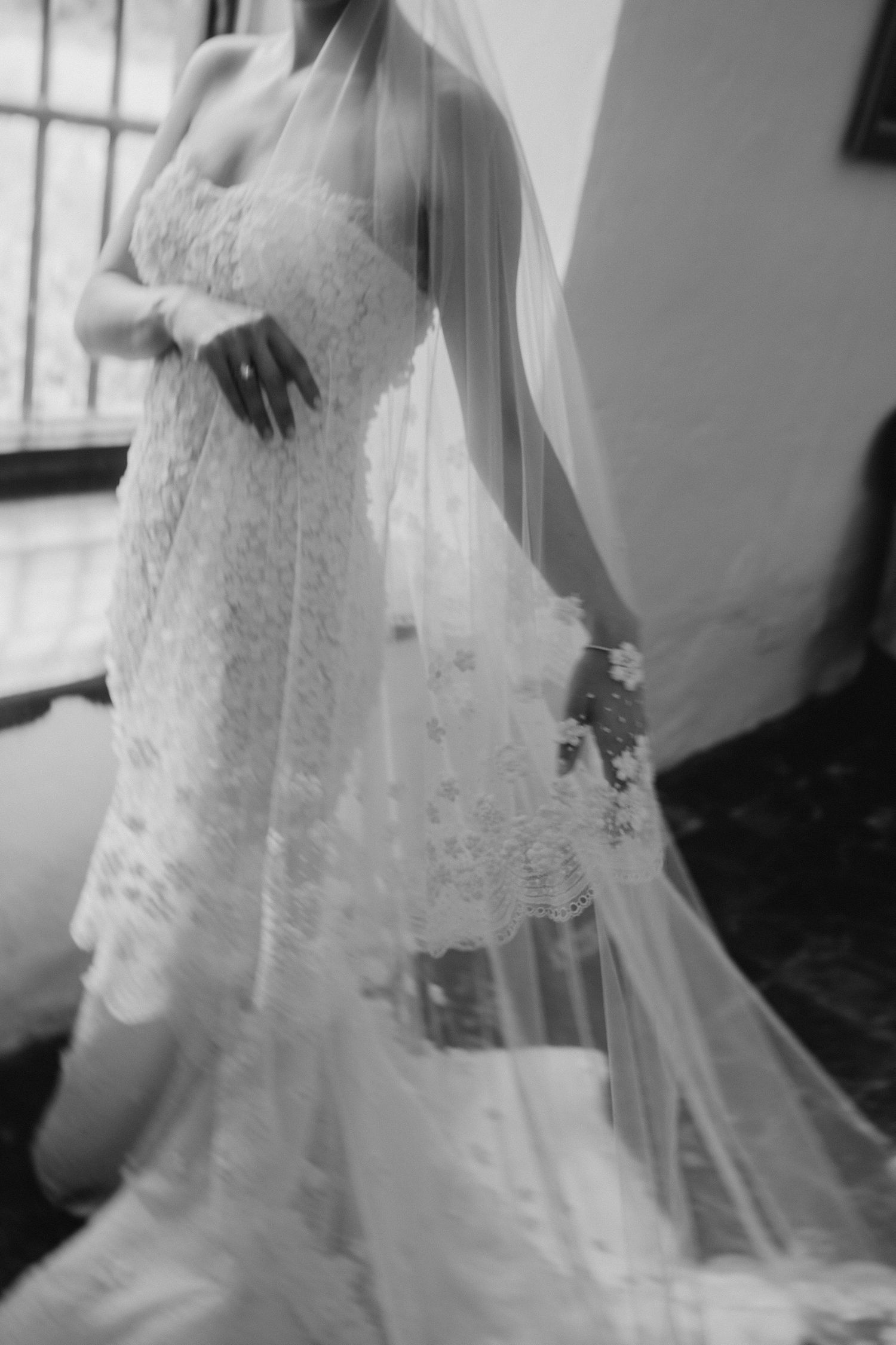Black and white photo of a bride in a lace wedding gown and flowing veil, holding a bouquet.