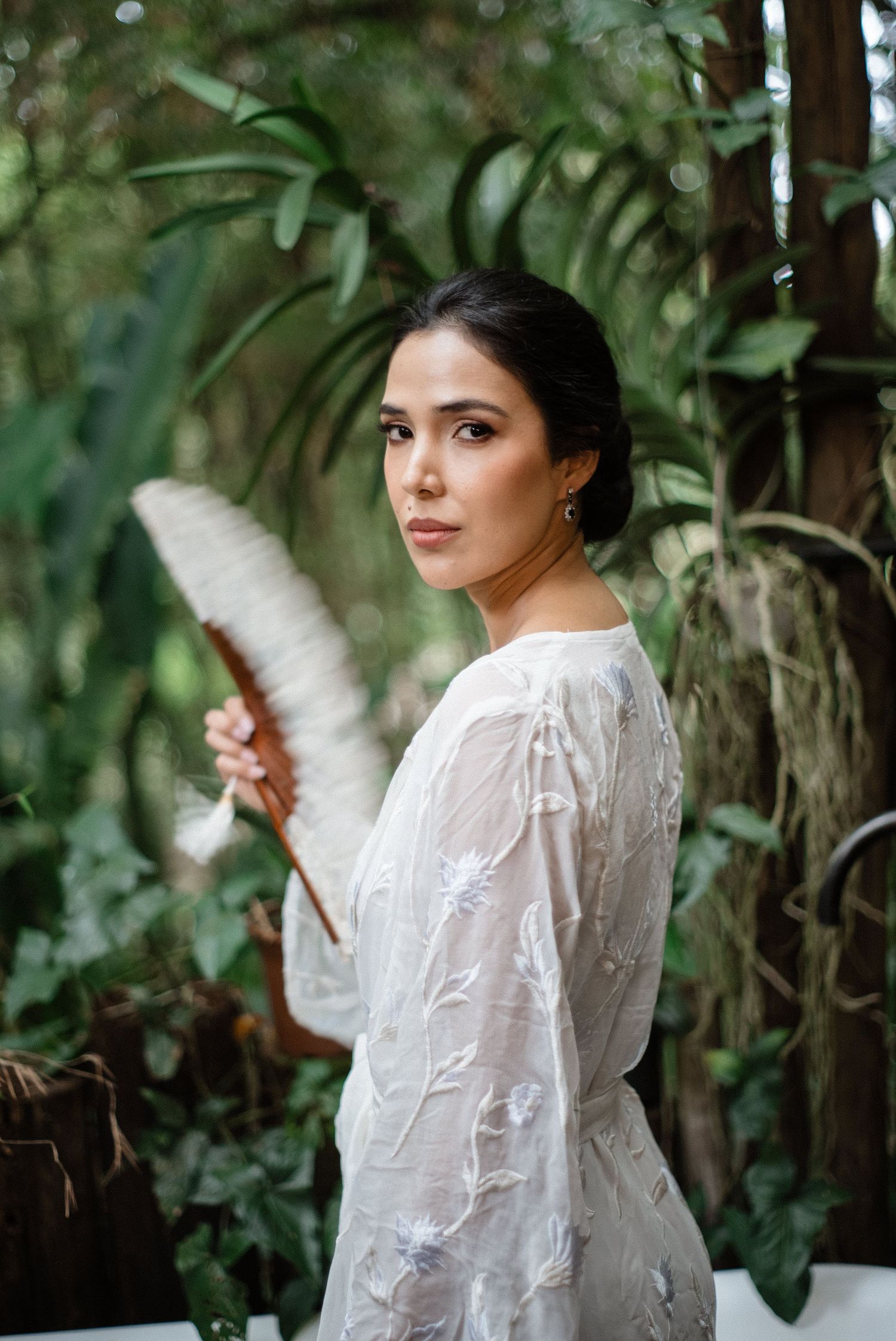 Dark-haired bride in sheer floral white dress holding a feather fan among tropical greenery.
