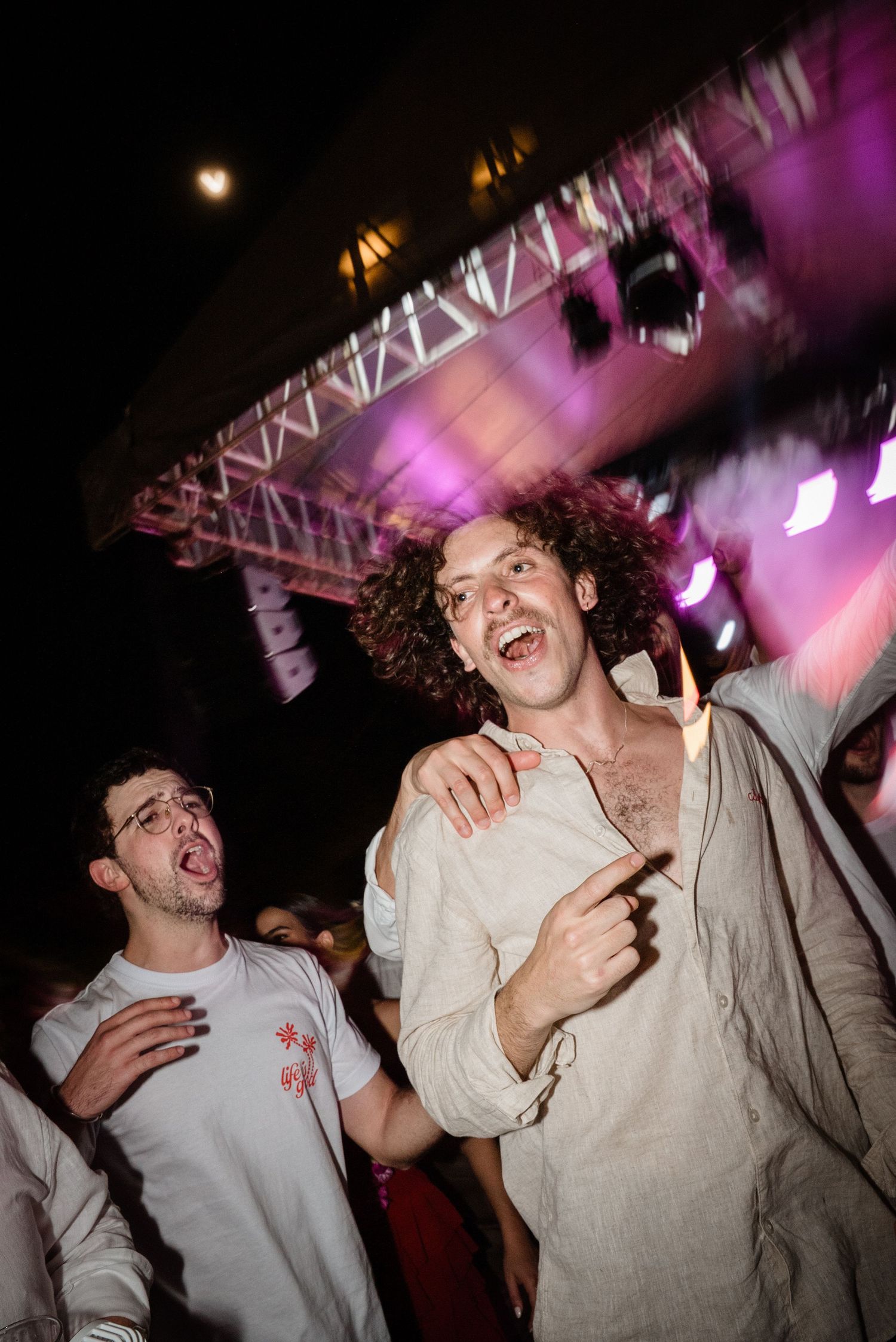 Excited curly-haired guest dancing enthusiastically at an outdoor wedding reception under colorful stage lights.