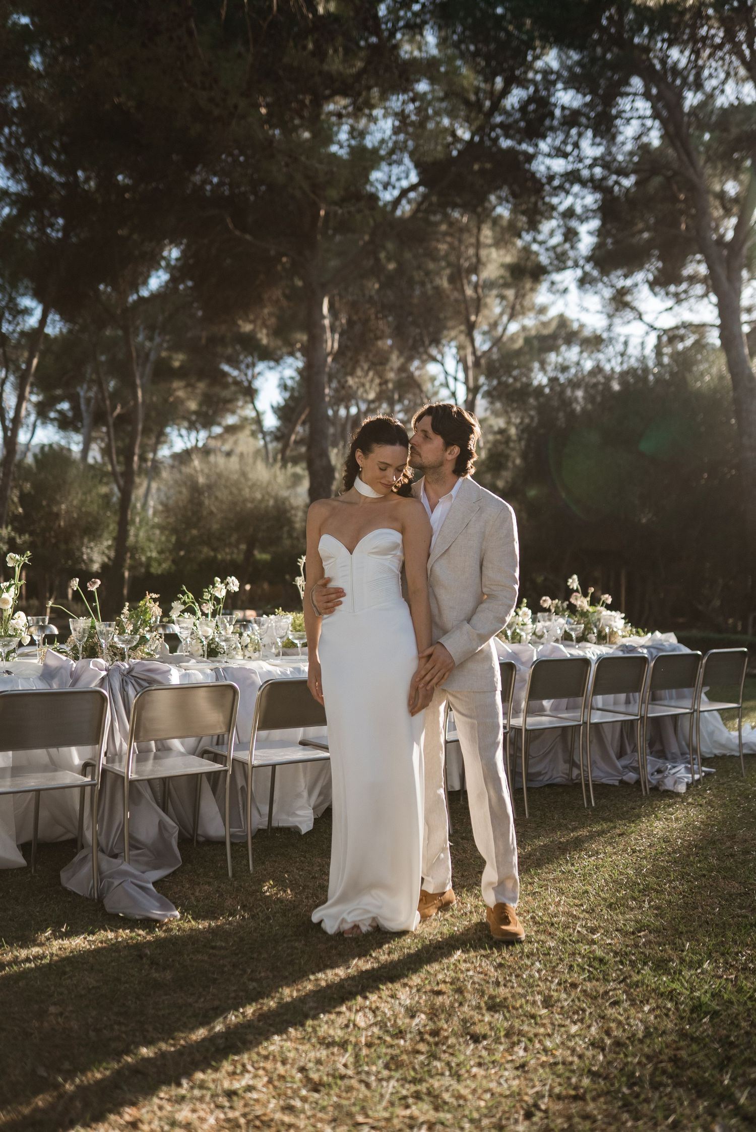 Bride and groom embrace outdoors beside elegant white reception tables set among tall pine trees in golden sunlight.