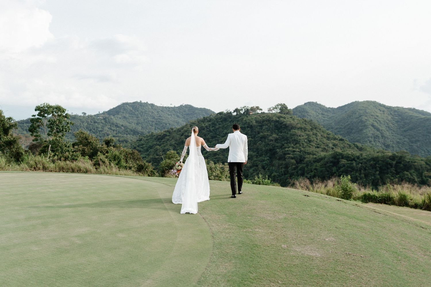 Bride and groom holding hands walking on lush green golf course with scenic mountain backdrop.