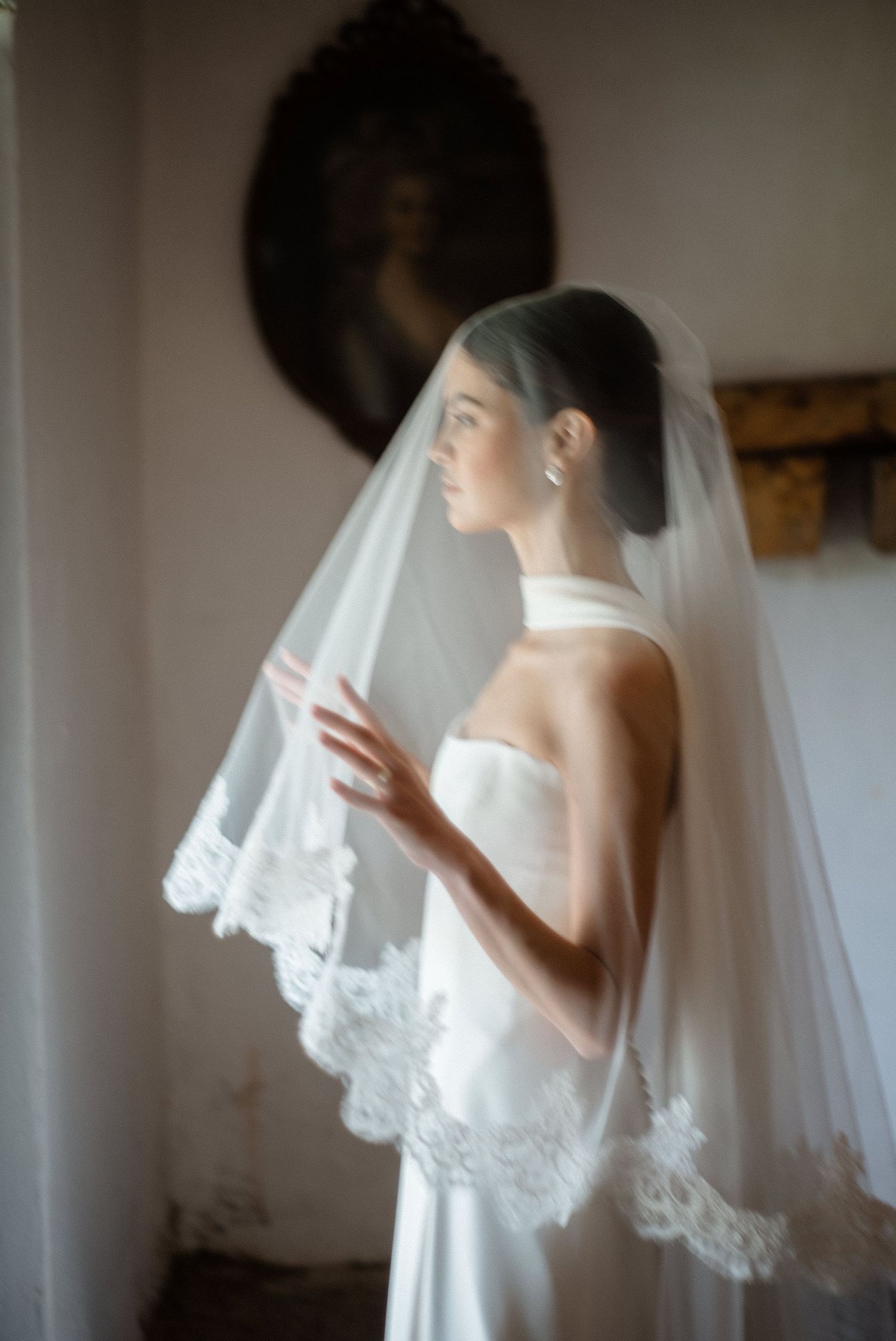 Dreamy soft-focus portrait of a bride in a halter gown holding a lace-trimmed veil in a vintage room.