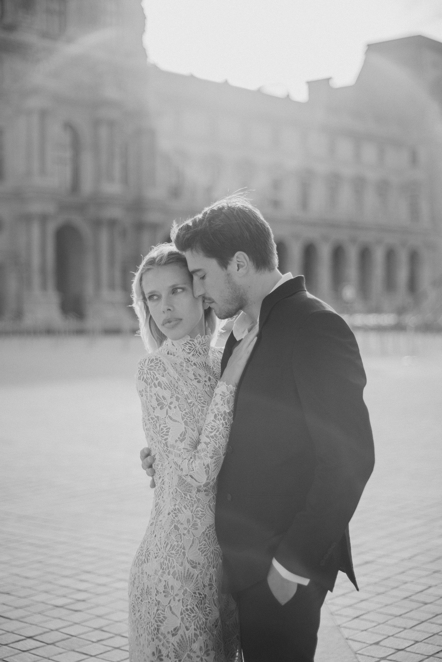 Black and white photo of an elegant couple in formal attire embracing near the Louvre in Paris.