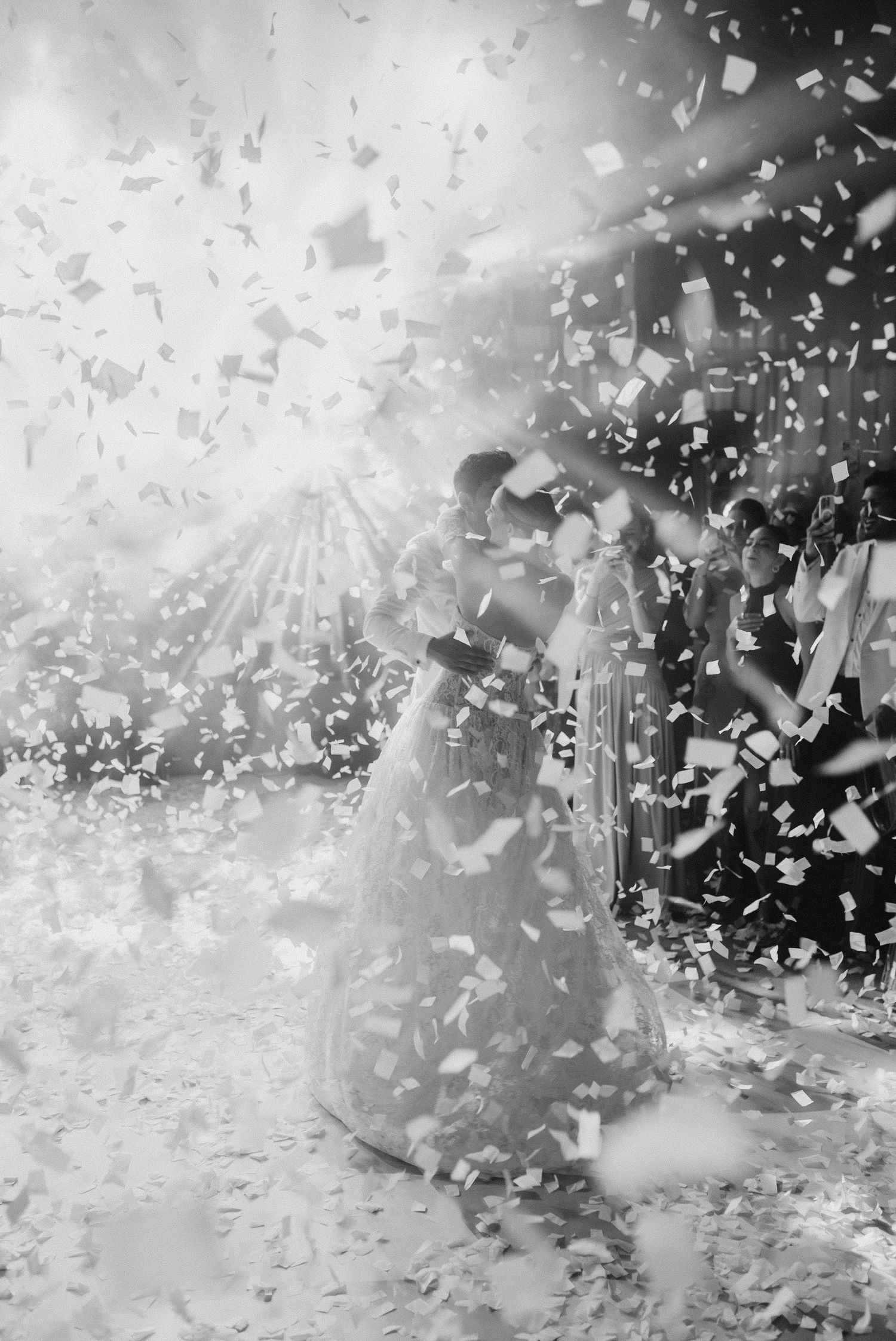 Bride and groom share first dance surrounded by falling confetti in a dramatic black and white wedding photo.