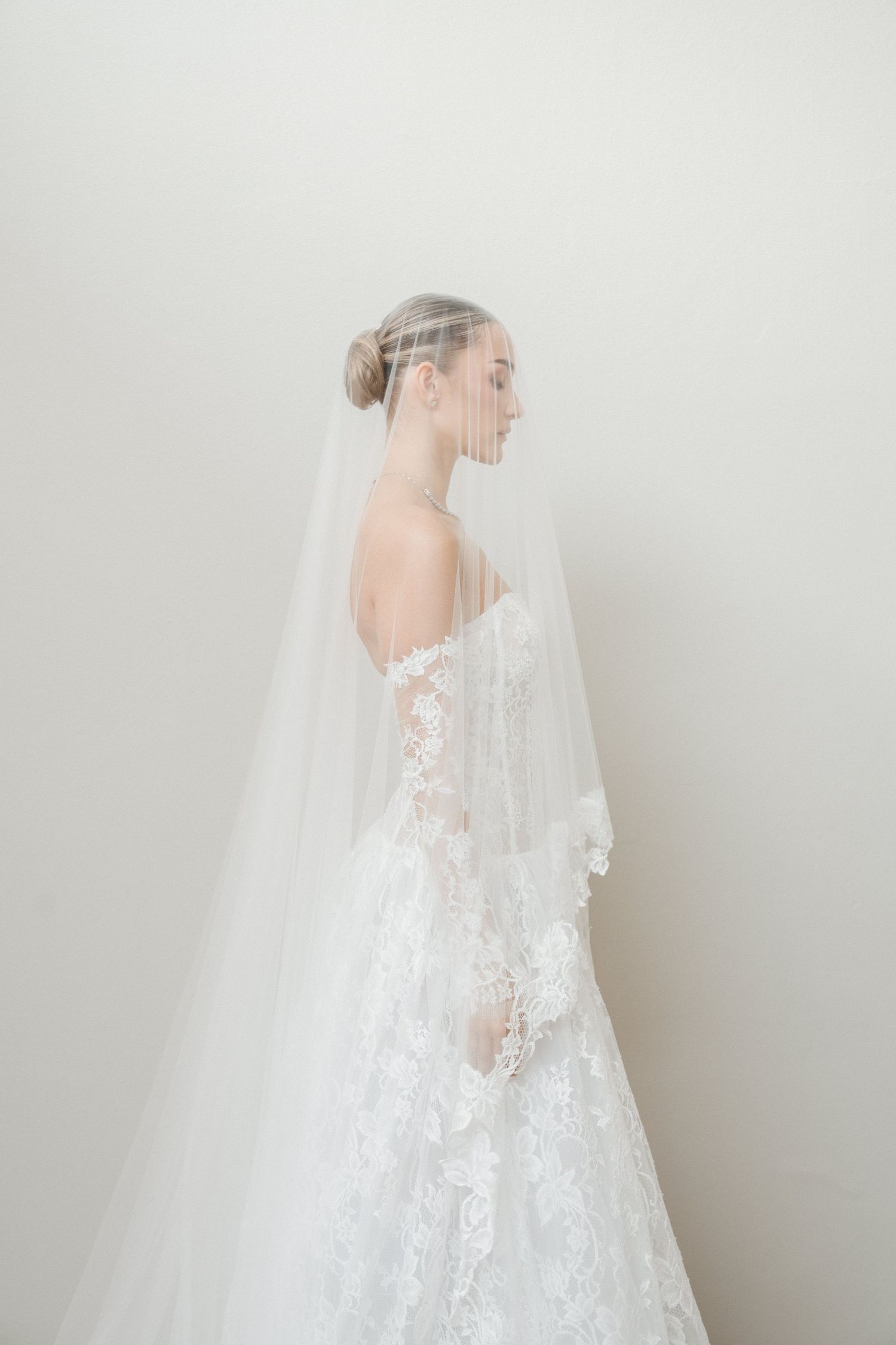 Elegant bride in lace gown with long cathedral veil poses against a white background.