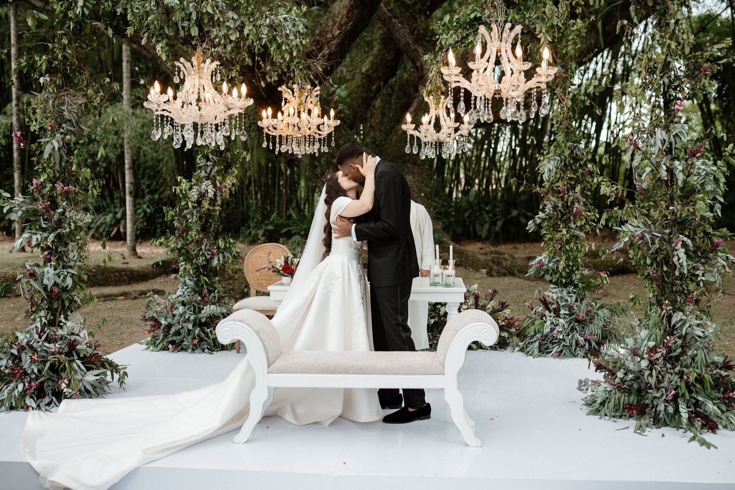 Bride and groom kiss beneath crystal chandeliers at elegant outdoor wedding ceremony.