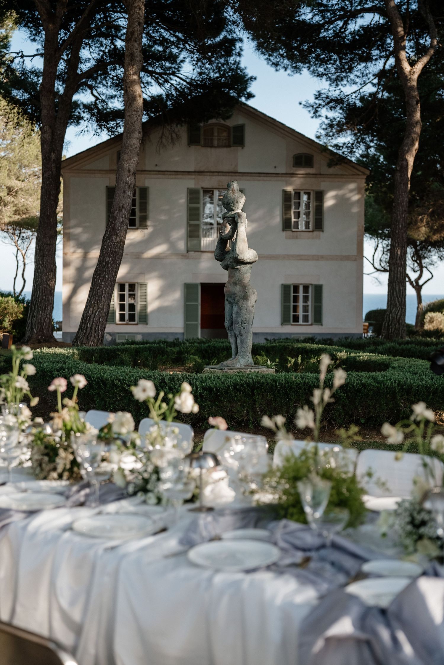 Elegant outdoor wedding reception tables with white linens before a classic villa featuring a bronze sculpture garden.