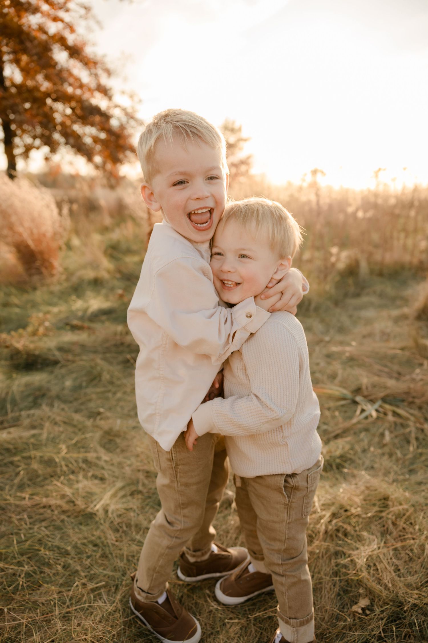 Two children in matching cream sweaters and khaki pants hugging and laughing together at sunset in a grassy field.