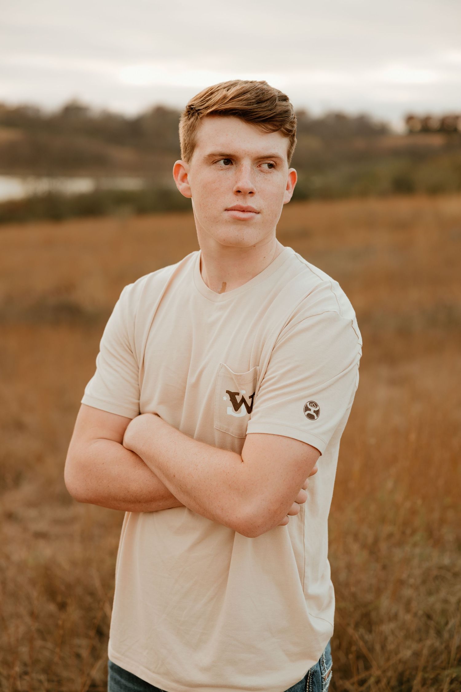 High school senior in a white t-shirt with arms crossed poses outdoors in a field at sunset.