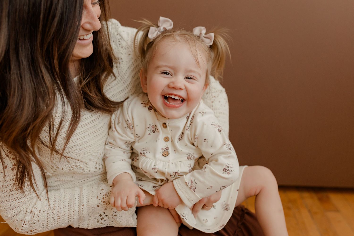 A joyful moment shared between parent and child sitting together on hardwood floor wearing cream-colored outfits.