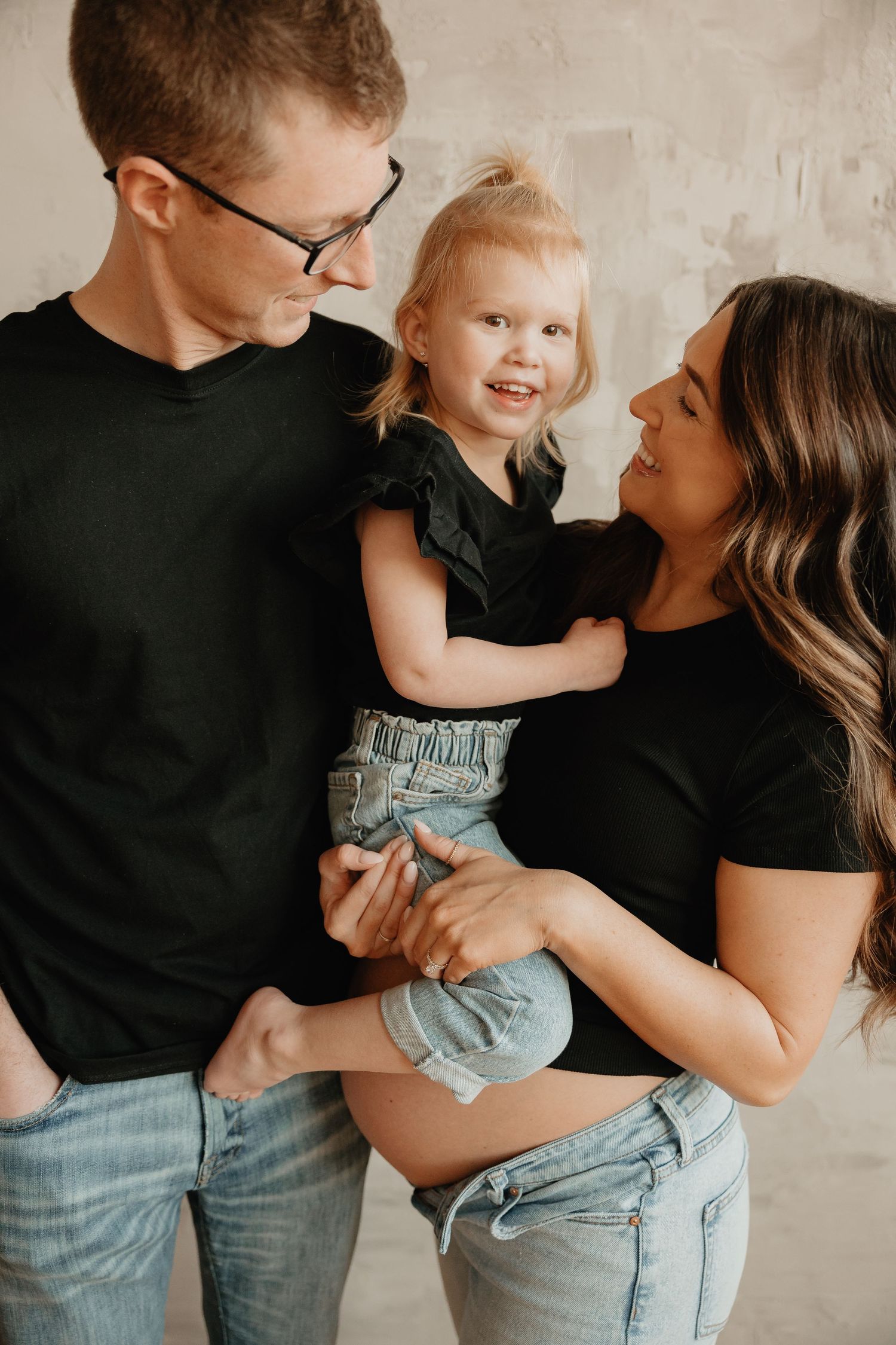 Family of three wearing black tops and jeans pose together against a neutral wall in a candid moment.