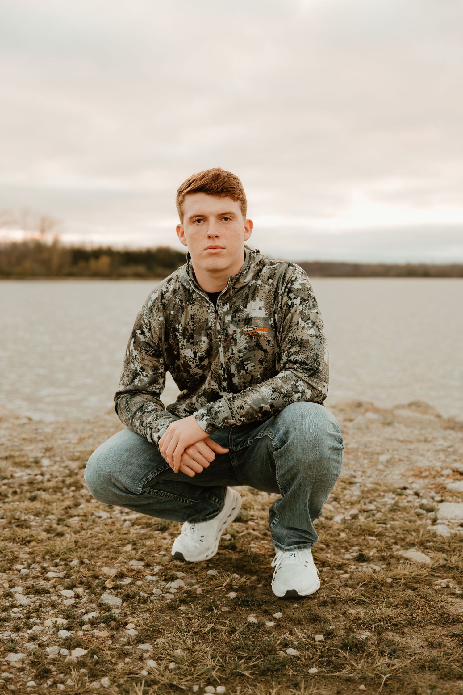 High school senior in a camouflage jacket and jeans crouches on a rocky shoreline near a lake at dusk.