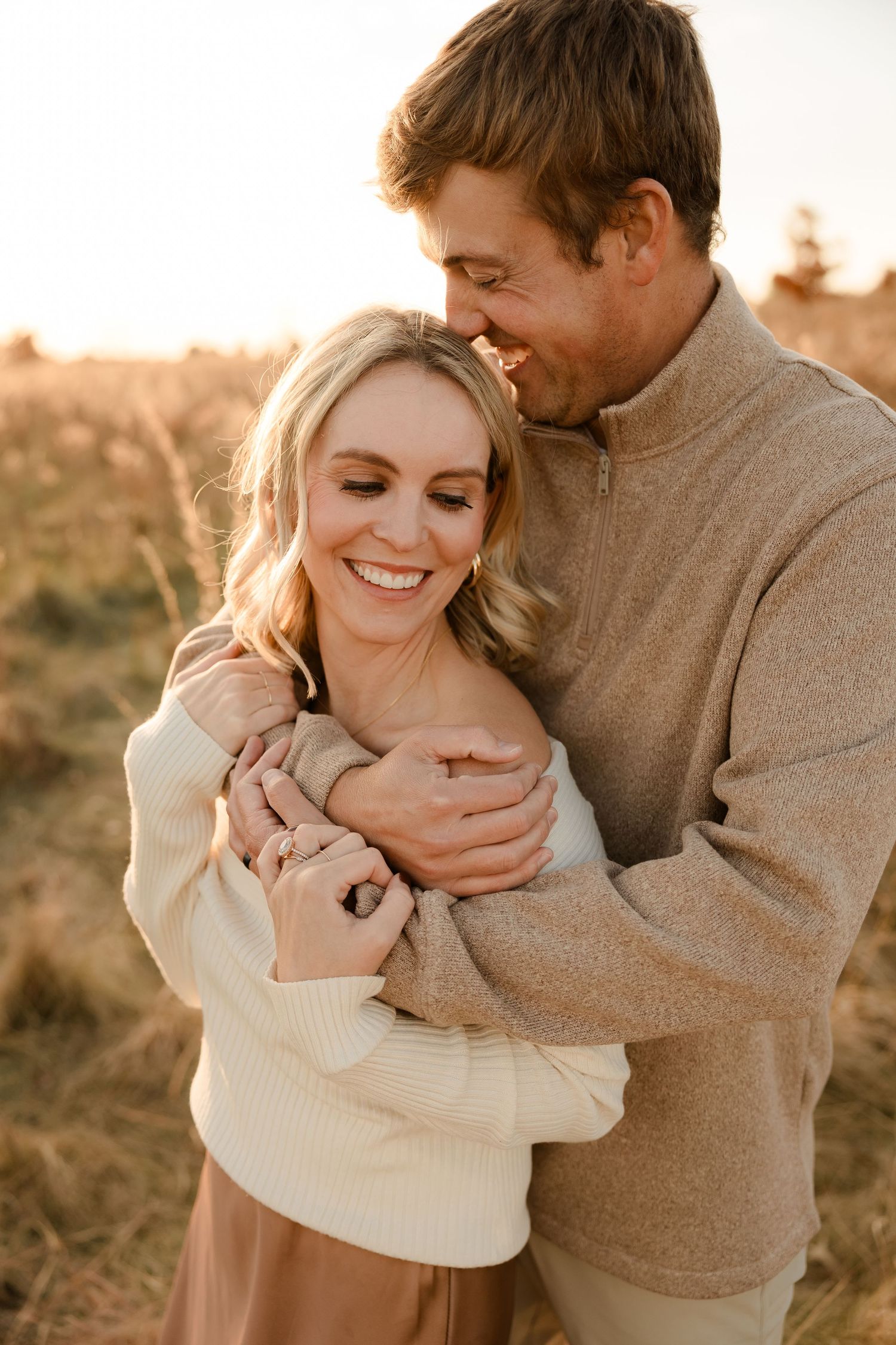 A loving couple embraces outdoors during golden hour wearing cozy sweaters and sharing a joyful moment together.