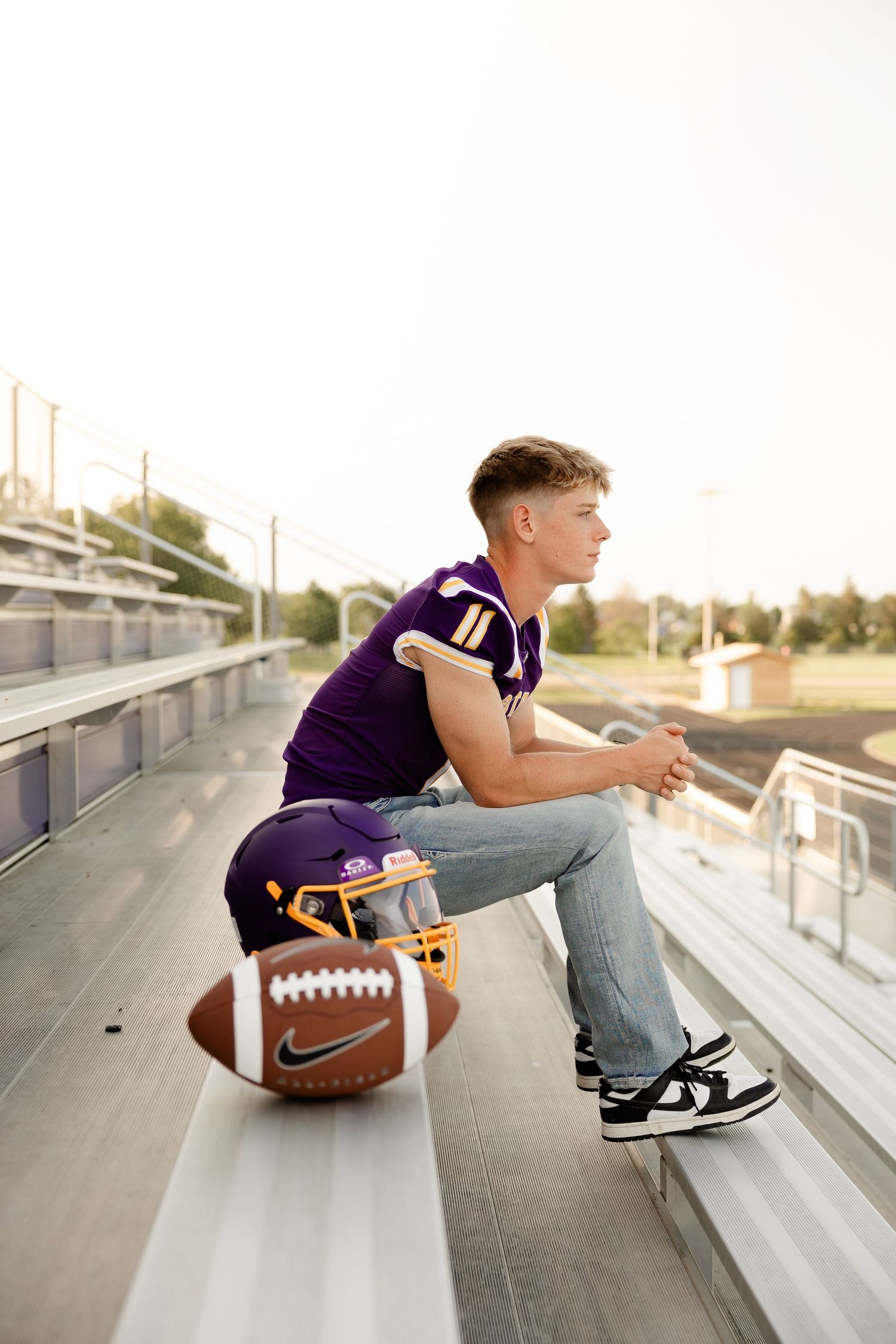 High school football player in purple jersey sits on stadium bleachers with football and equipment beside him.