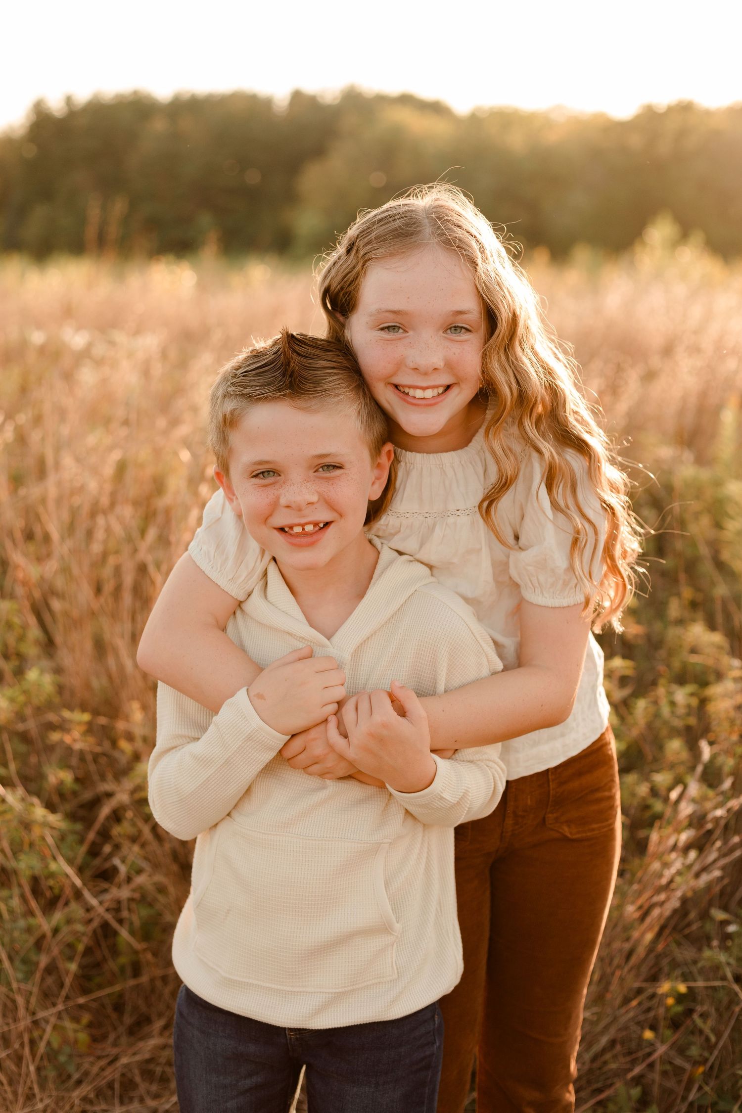 Two children embrace and laugh together in a golden sunset field with tall grass and warm lighting.
