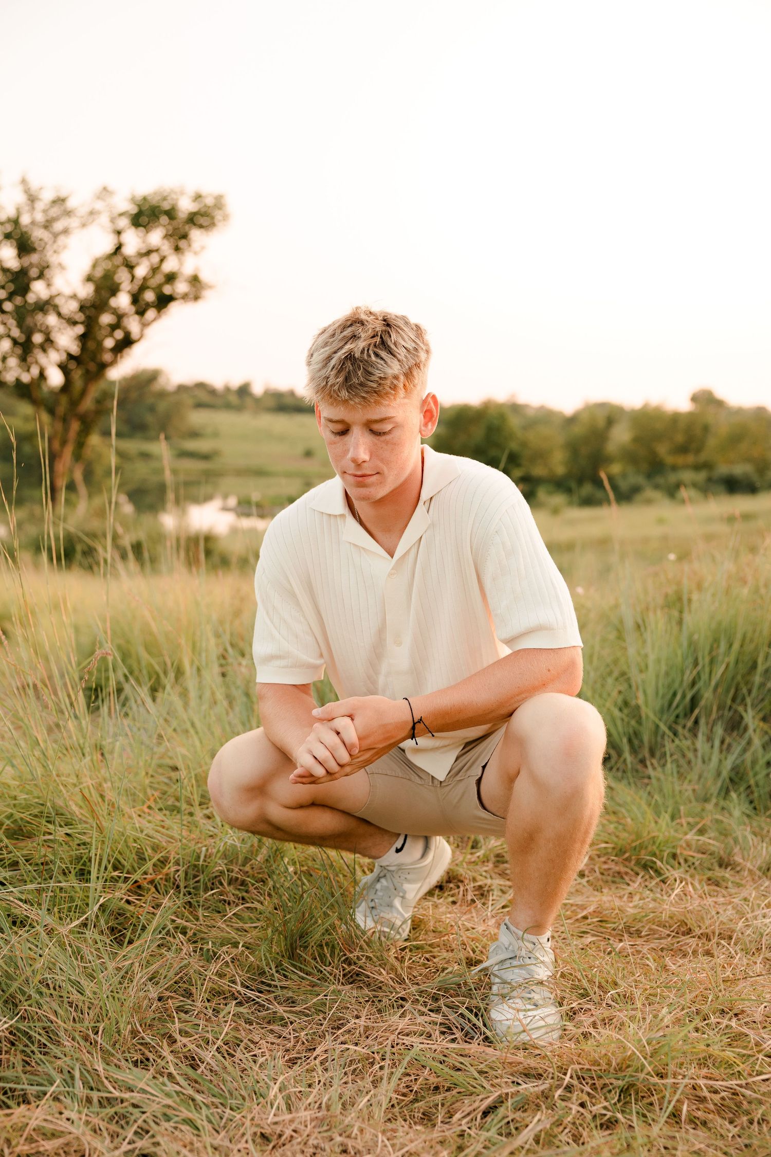 A high school senior in a white polo shirt and khaki shorts crouches in a grassy meadow at sunset.