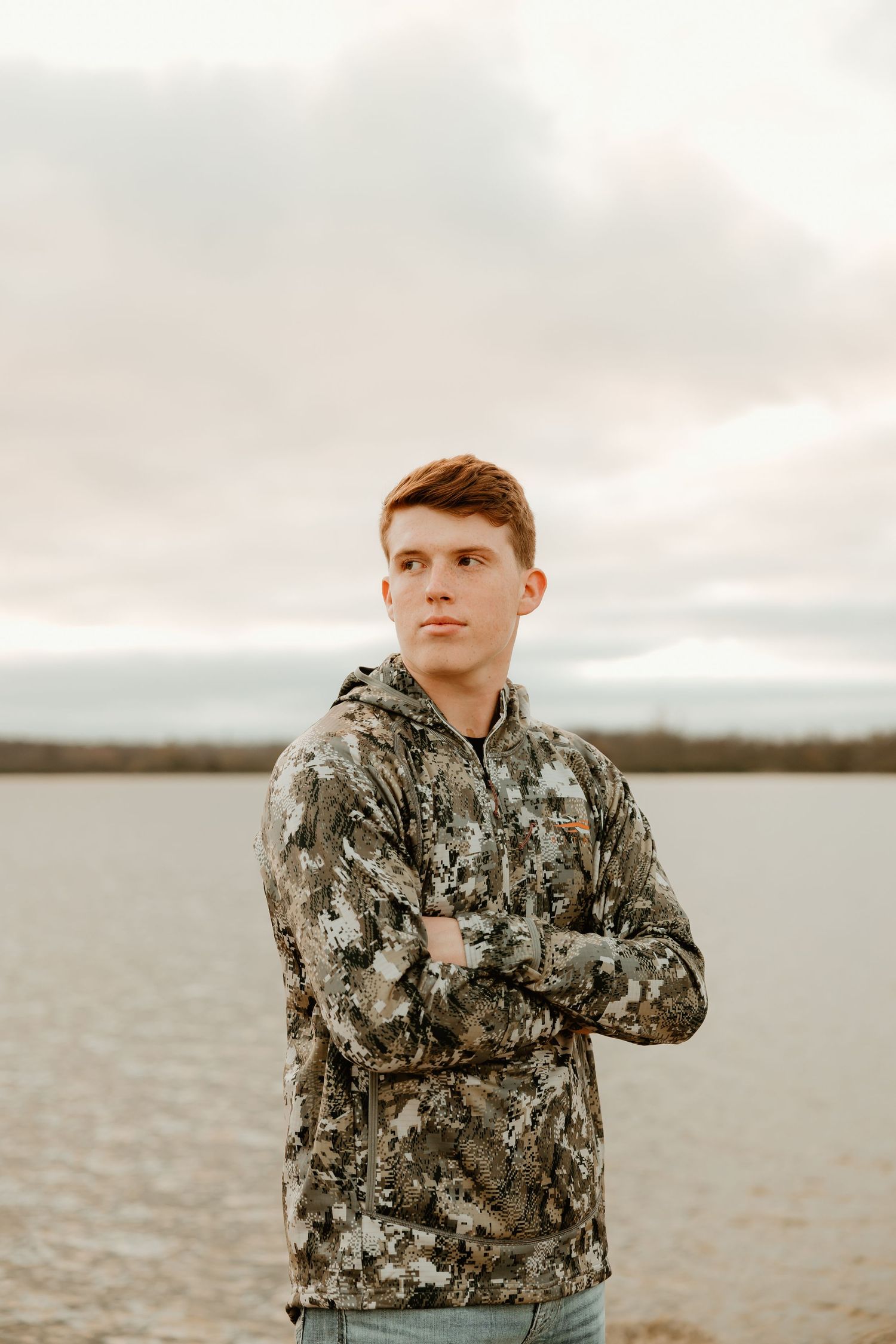 High school senior wearing a camouflage hoodie stands with arms crossed by a lake during overcast weather.