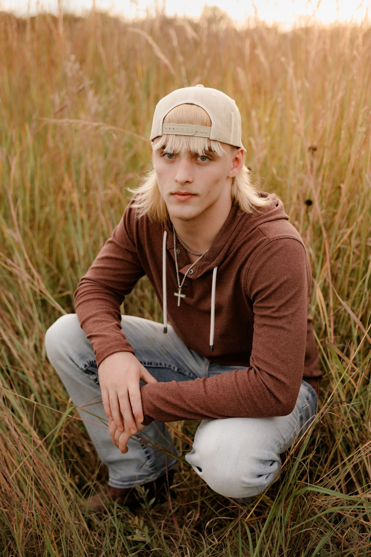 A high school senior in a brown hoodie and baseball cap crouches in tall golden grass during sunset.