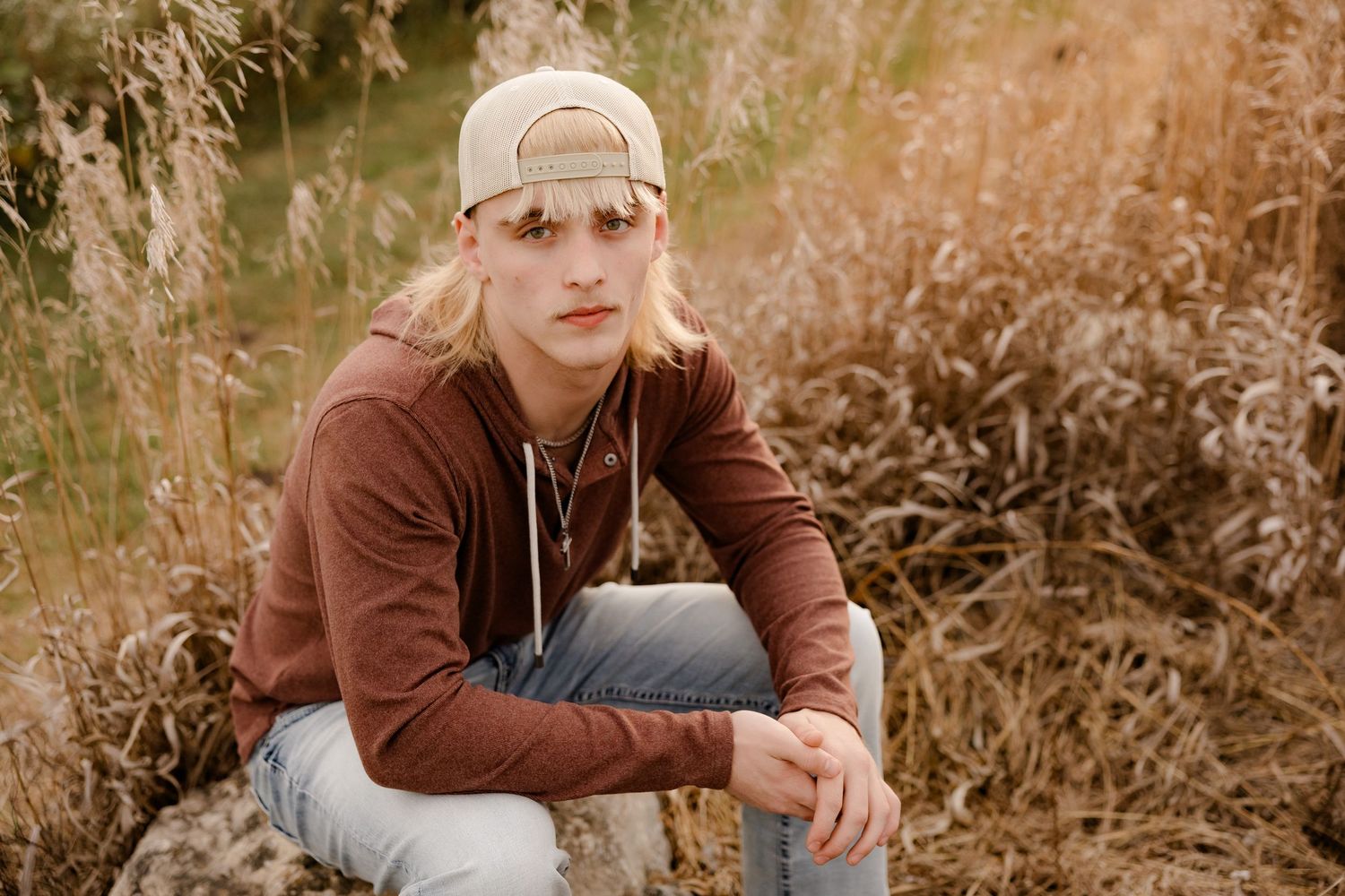 High school senior in brown sweater and backwards cap sits thoughtfully among tall dry grass in golden outdoor setting.