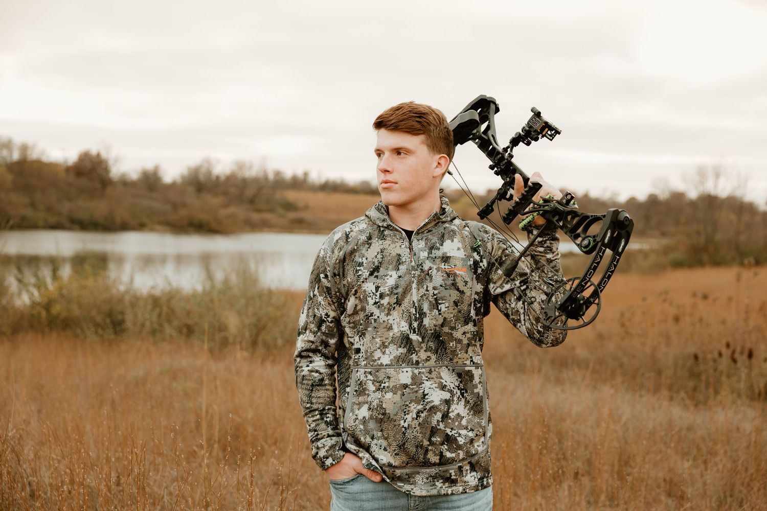 High school senior in camouflage gear stands in autumn field holding compound bow with lake and cloudy sky in background.