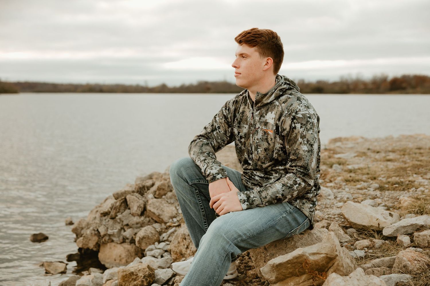 High school senior in camouflage jacket and jeans sits on rocky shore beside lake on cloudy day.