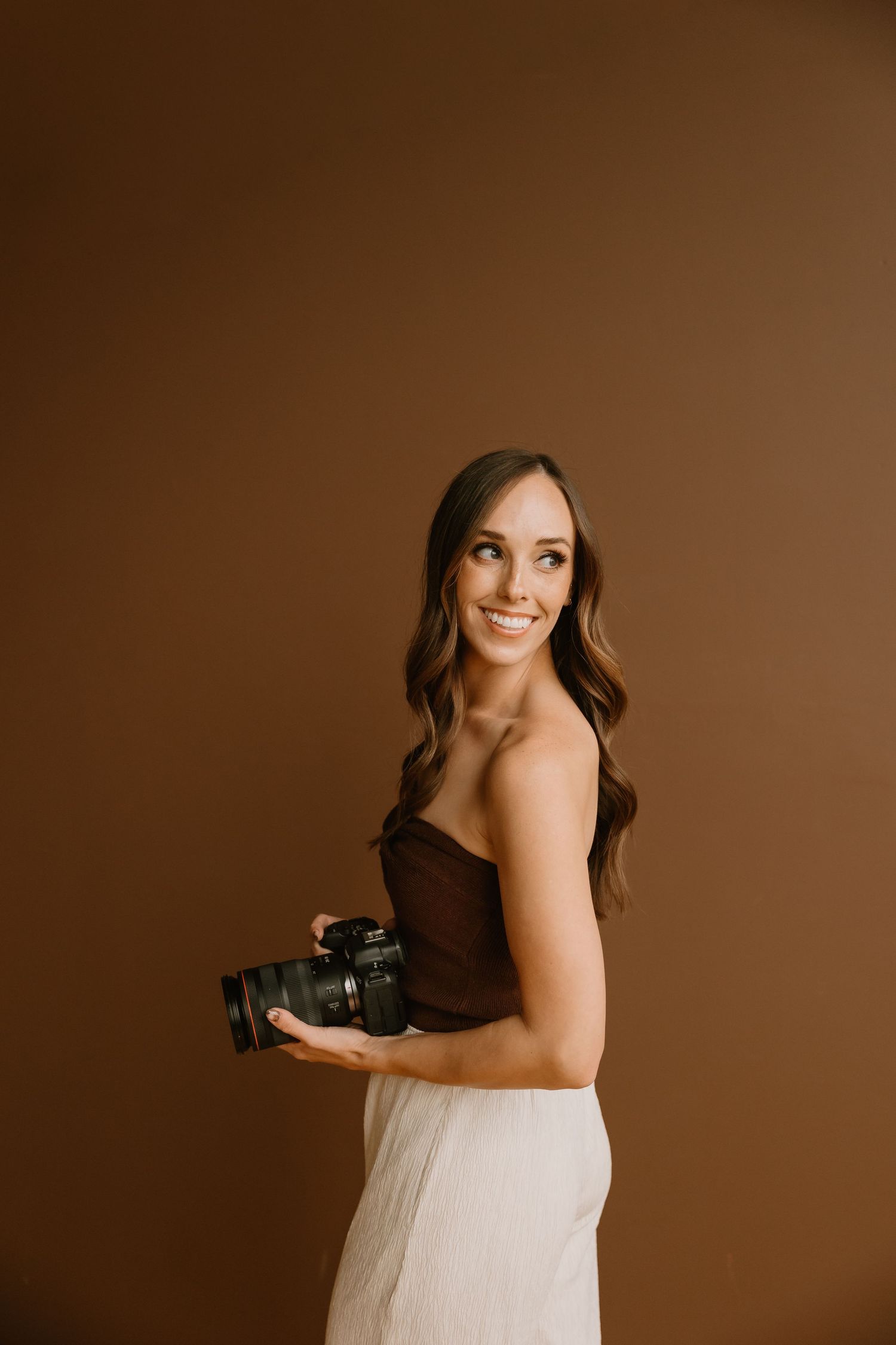 Professional photographer in a brown strapless top and white pants holds a mirrorless camera against brown backdrop.