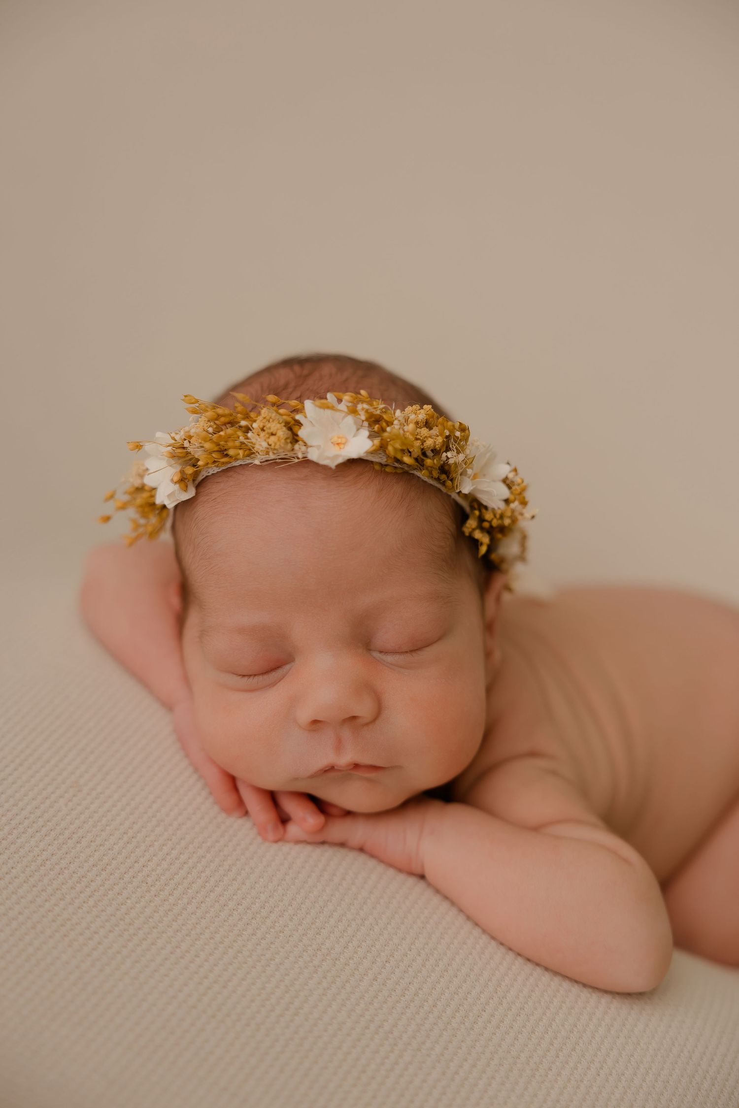 Peacefully sleeping newborn wearing delicate floral crown rests on cream-colored fabric in soft lighting.