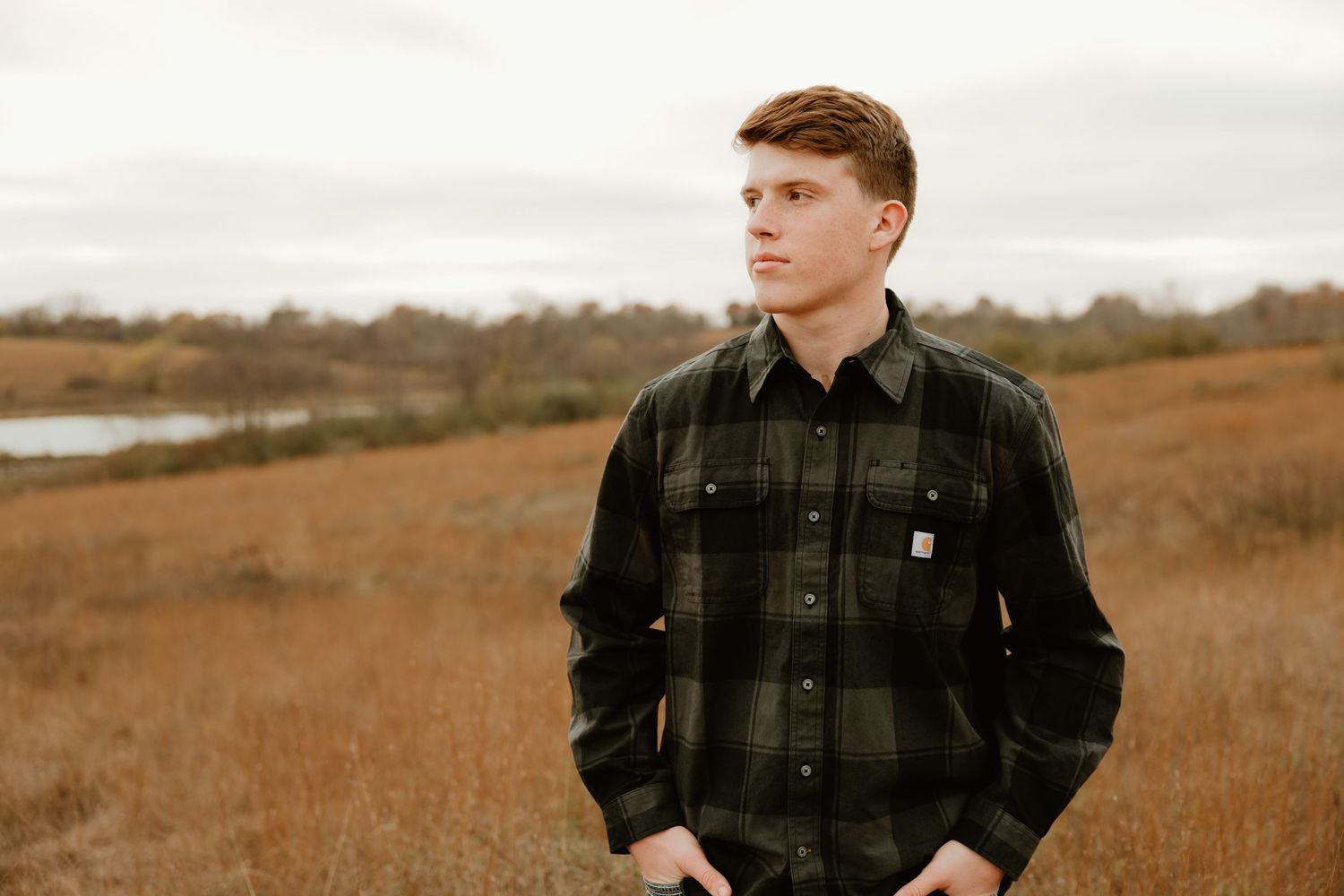 High school senior wearing black plaid jacket stands thoughtfully in autumn field during golden hour with nature in background.
