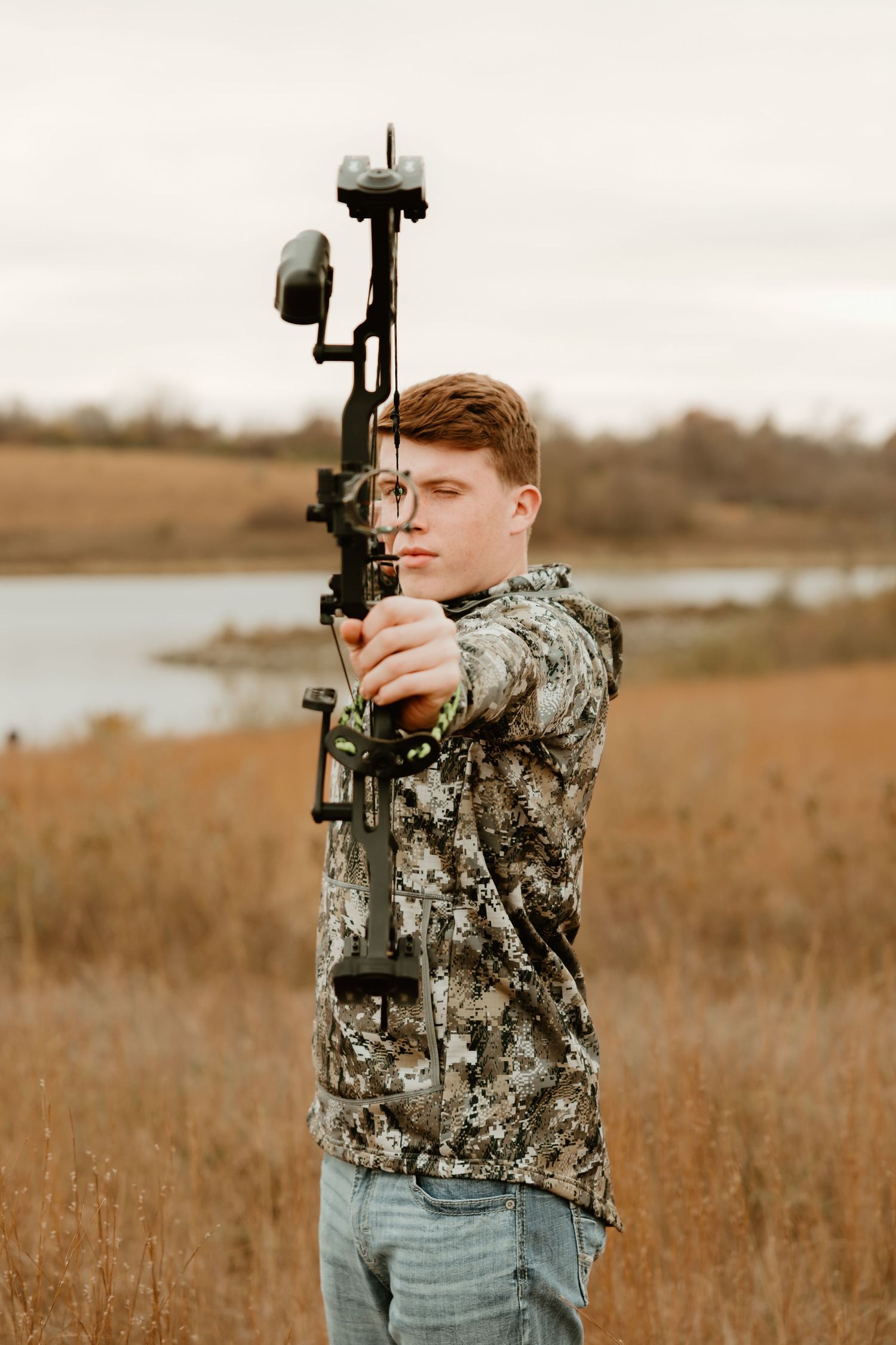 A high school senior in camouflage clothing aims a compound bow in a grassy field near a lake.
