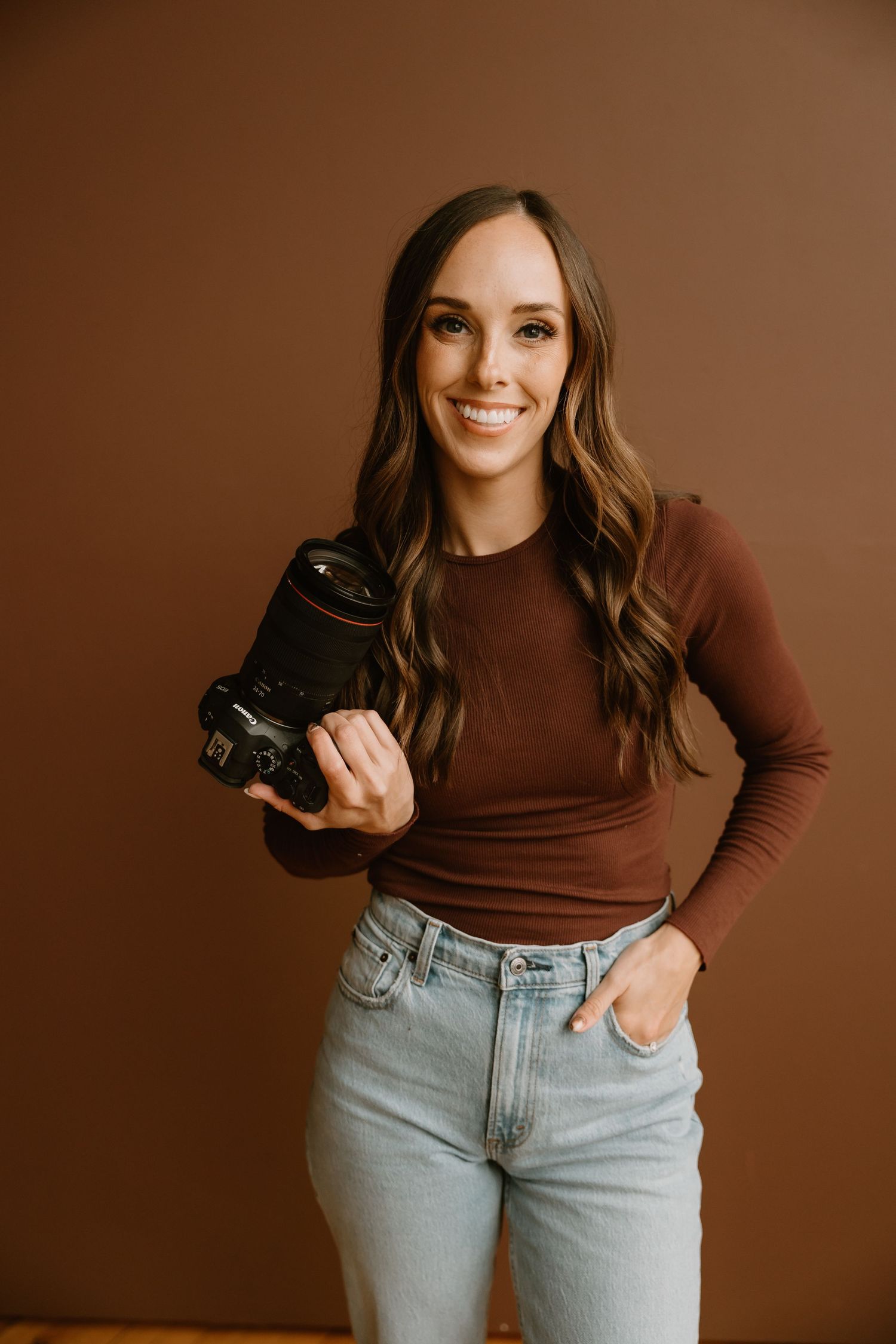 Professional photographer in brown top and light denim poses with mirrorless camera against brown backdrop.