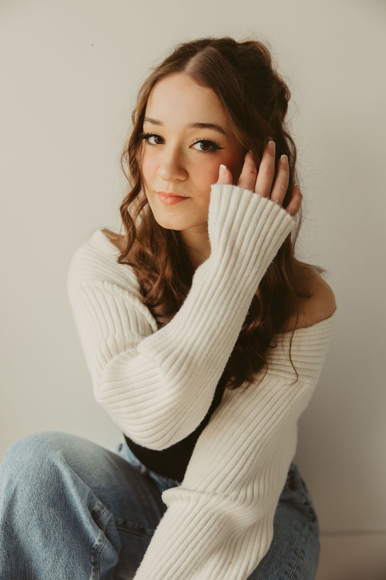 Portrait in cozy white knit sweater and jeans against neutral wall, with long wavy brown hair and natural makeup.